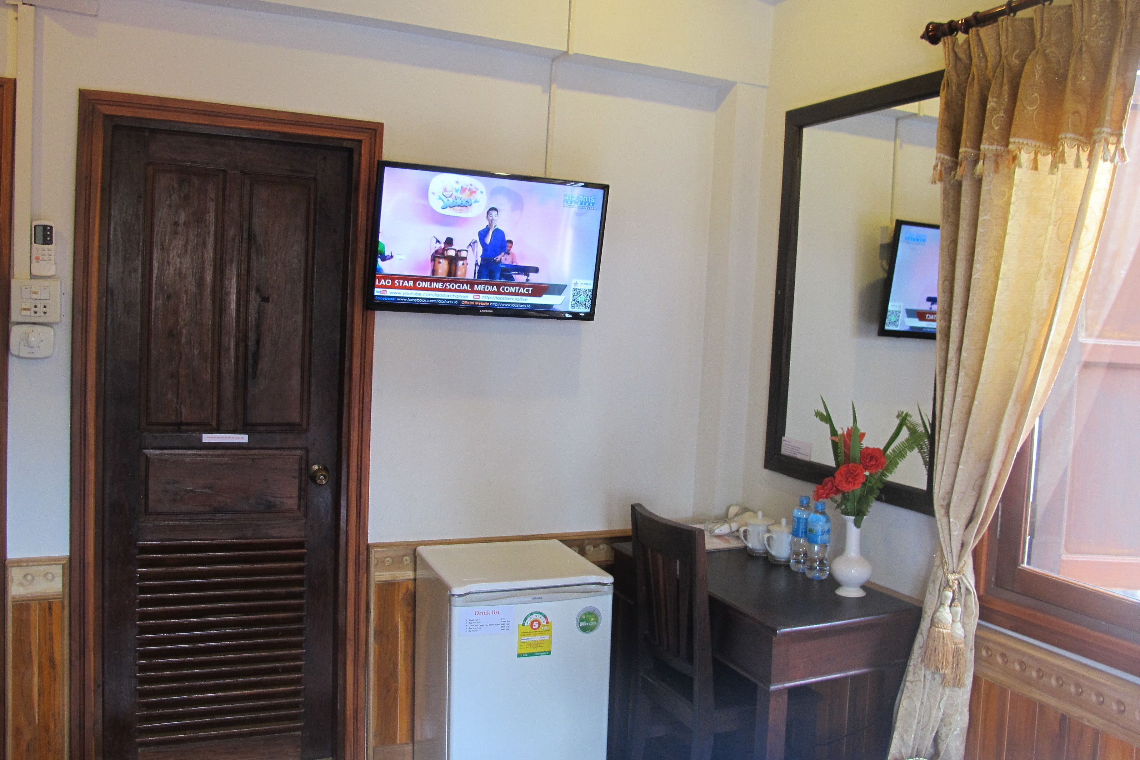 Una camera d'albergo con una porta in legno, un frigorifero bianco, una televisione montata a parete, un tavolo di legno e uno specchio con cornice decorativa.