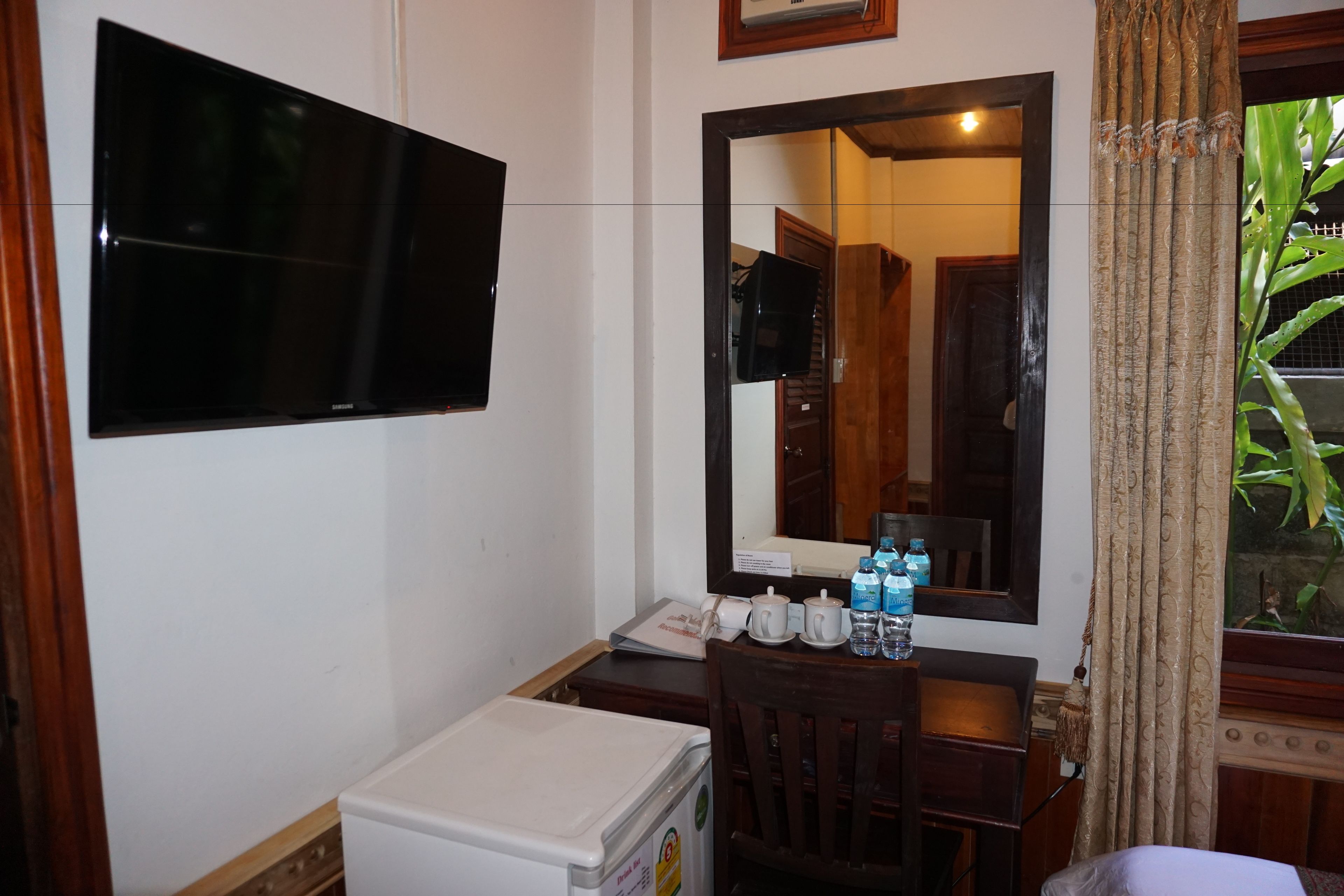 A small room with a flat-screen TV mounted on the wall, a wooden desk with a chair, a small refrigerator, and a window with curtains.