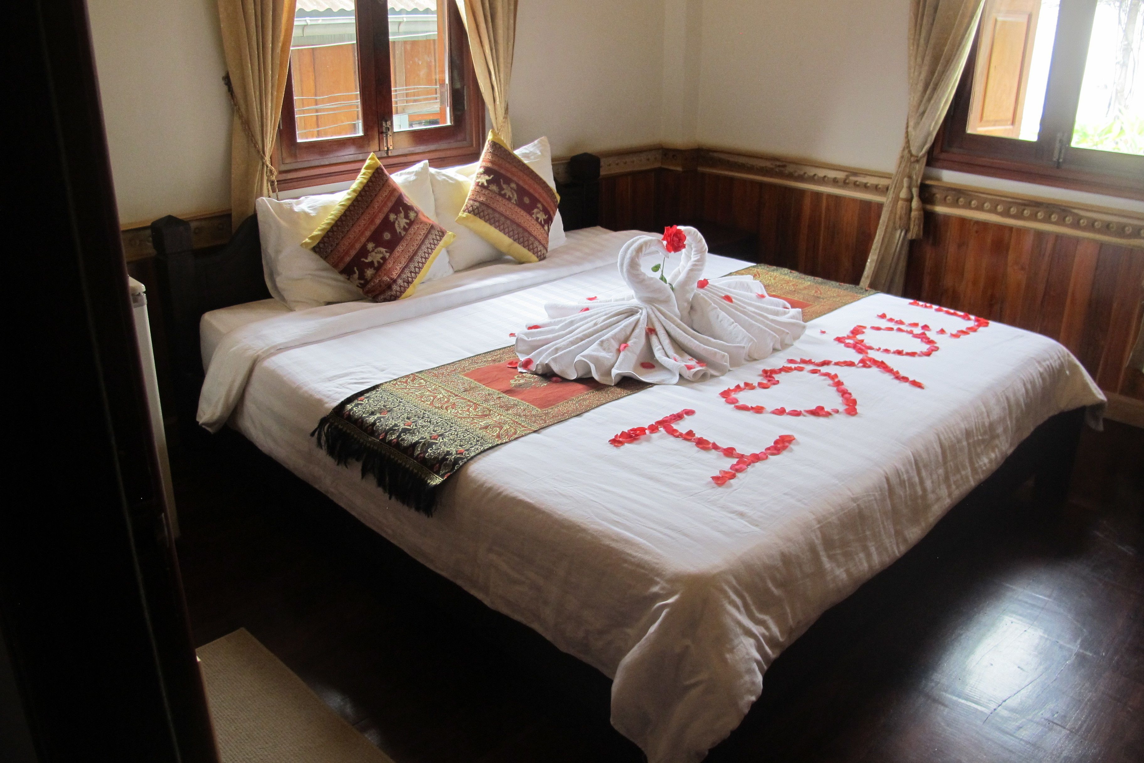 A neatly made bed with a white bedspread, a decorative swan towel, and a heart-shaped arrangement of rose petals spelling "I love you".