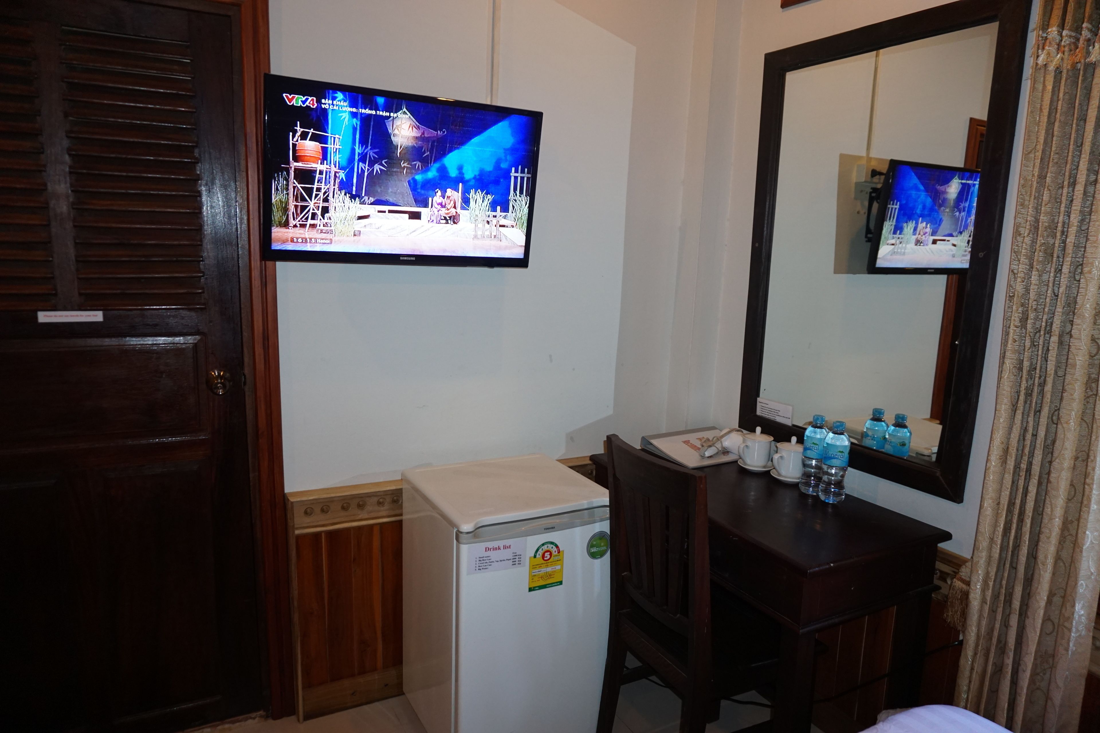 Habitación de hotel con televisor montado en la pared, un refrigerador pequeño, un escritorio de madera con espejo y una puerta con marco de madera.