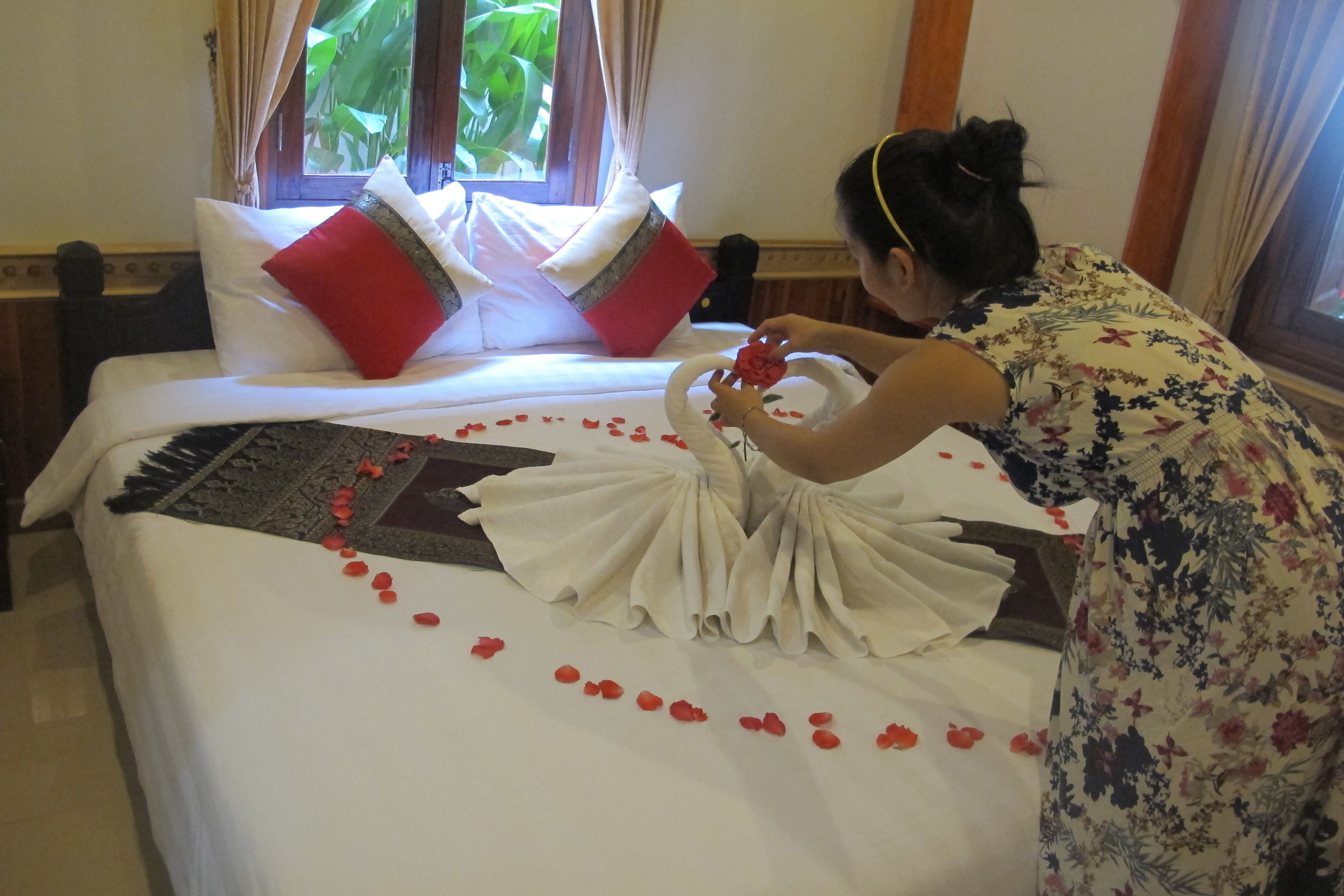 A bed with red rose petals arranged on it, a white bedspread, and a woman arranging a flower.