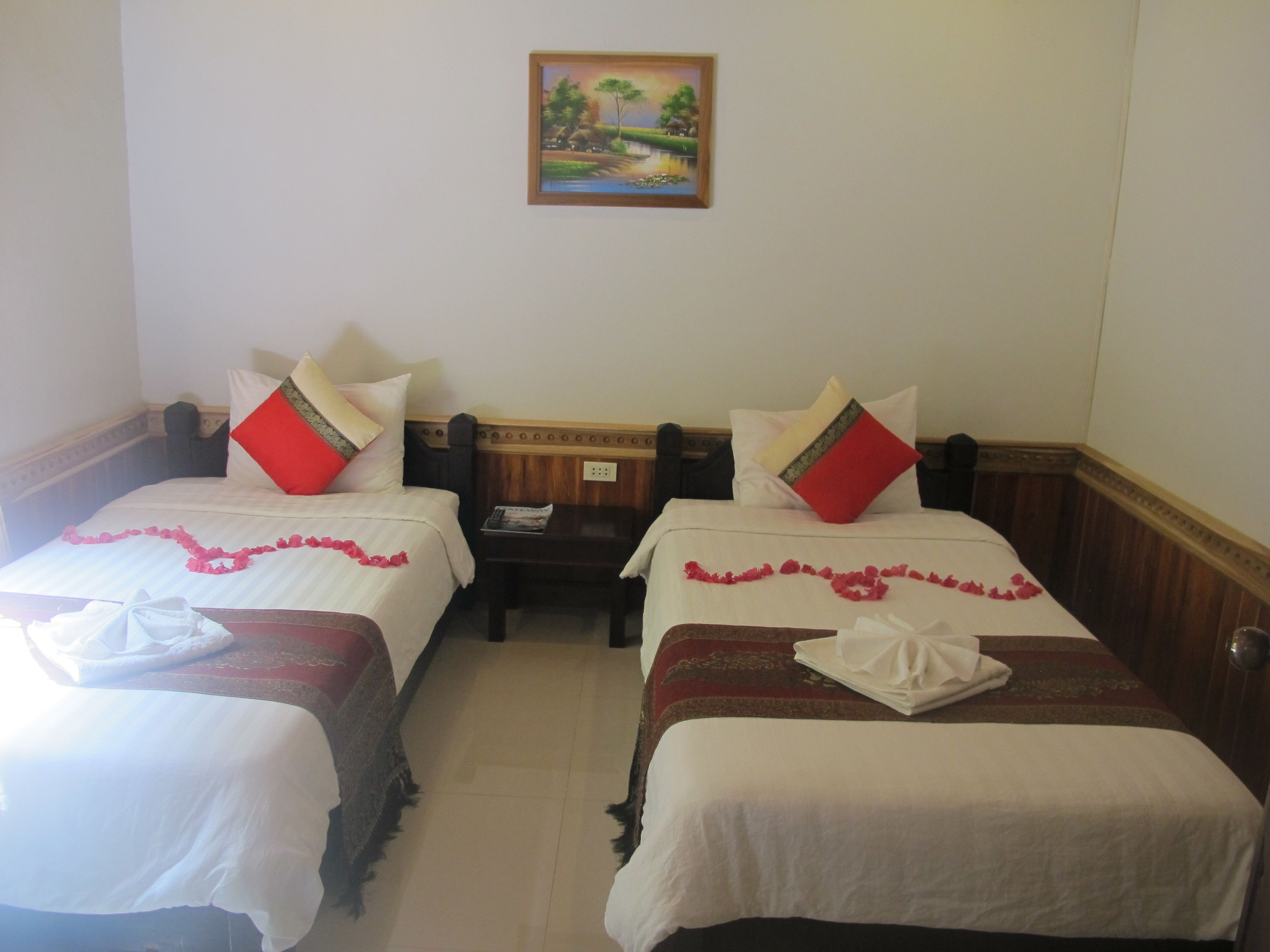Een hotelkamer met twee bedden, elk met een rood-wit kussen, een houten hoofdbord en een schilderij aan de muur.