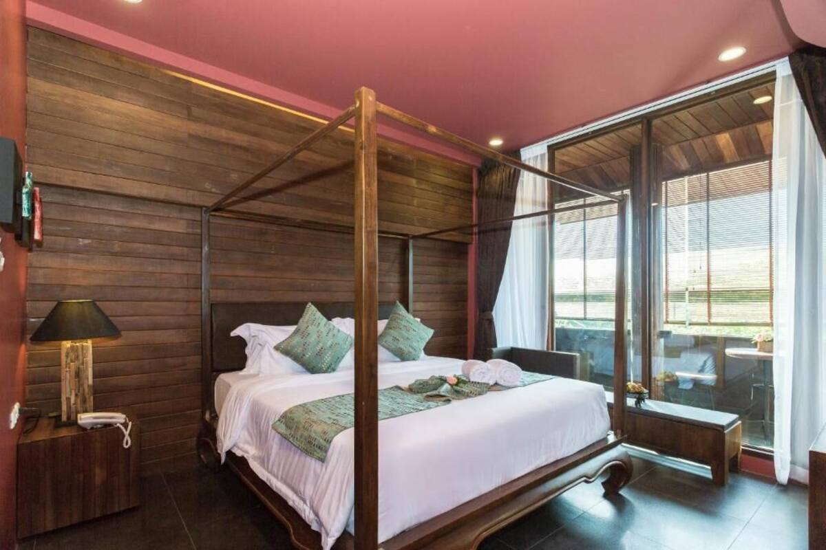 A four-poster bed with a wooden headboard, a bedside table with a lamp, and a view of the outdoors through large windows.