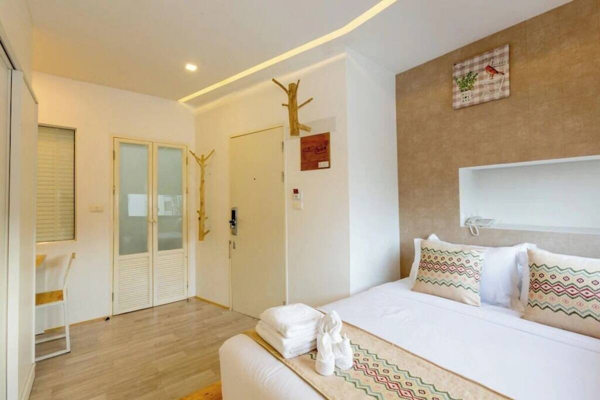A modern hotel room with a bed, a small seating area, a door, and a wall-mounted shelf.