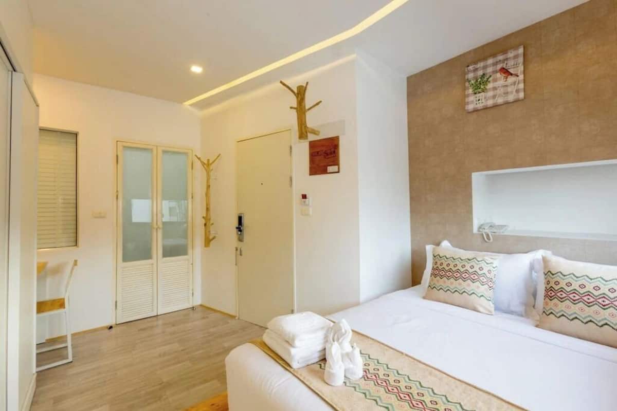 A modern hotel room with a bed, a small seating area, a door, and a wall-mounted shelf.