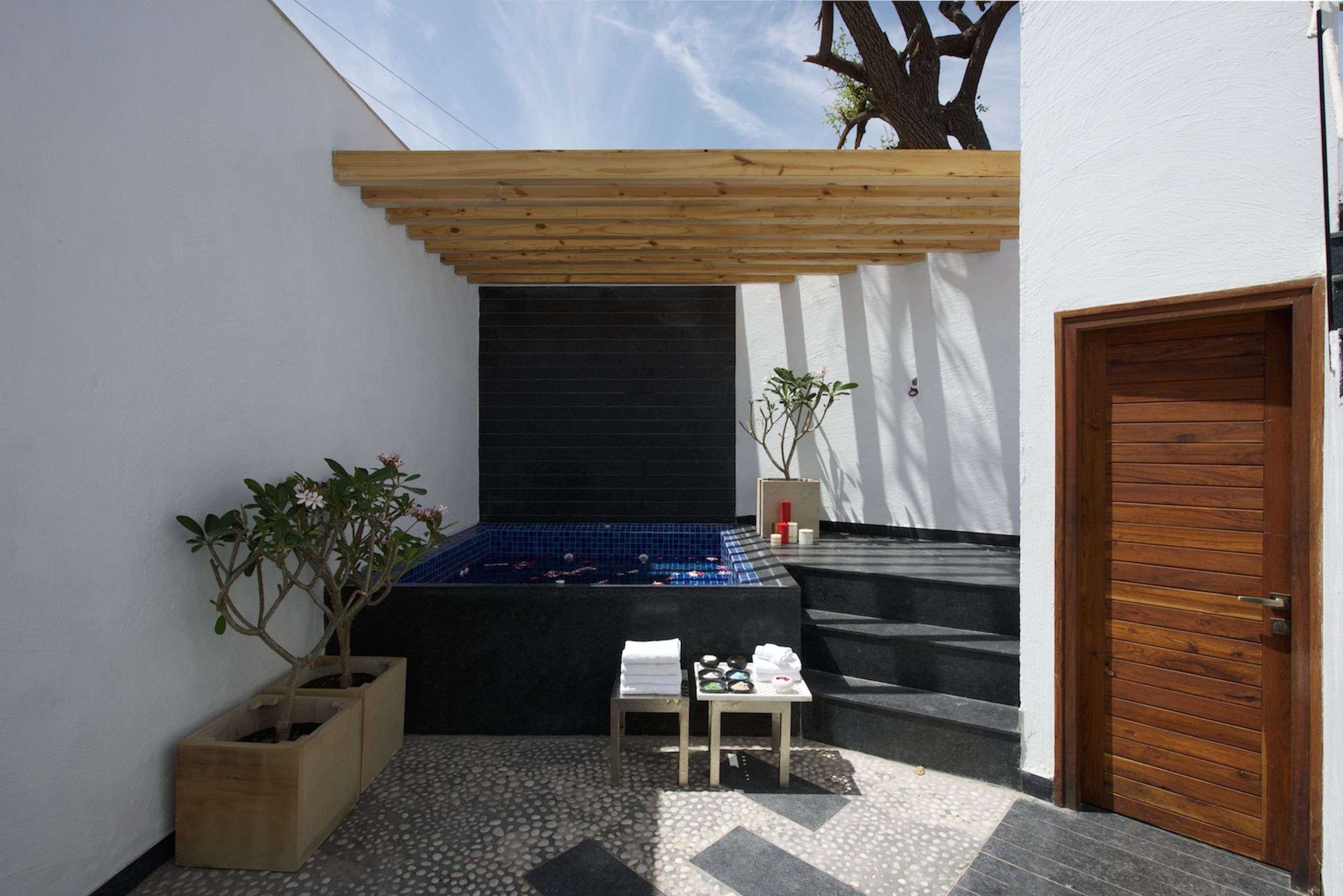 A spa area with a wooden pergola, a black wall, a wooden door, and a small table with towels.