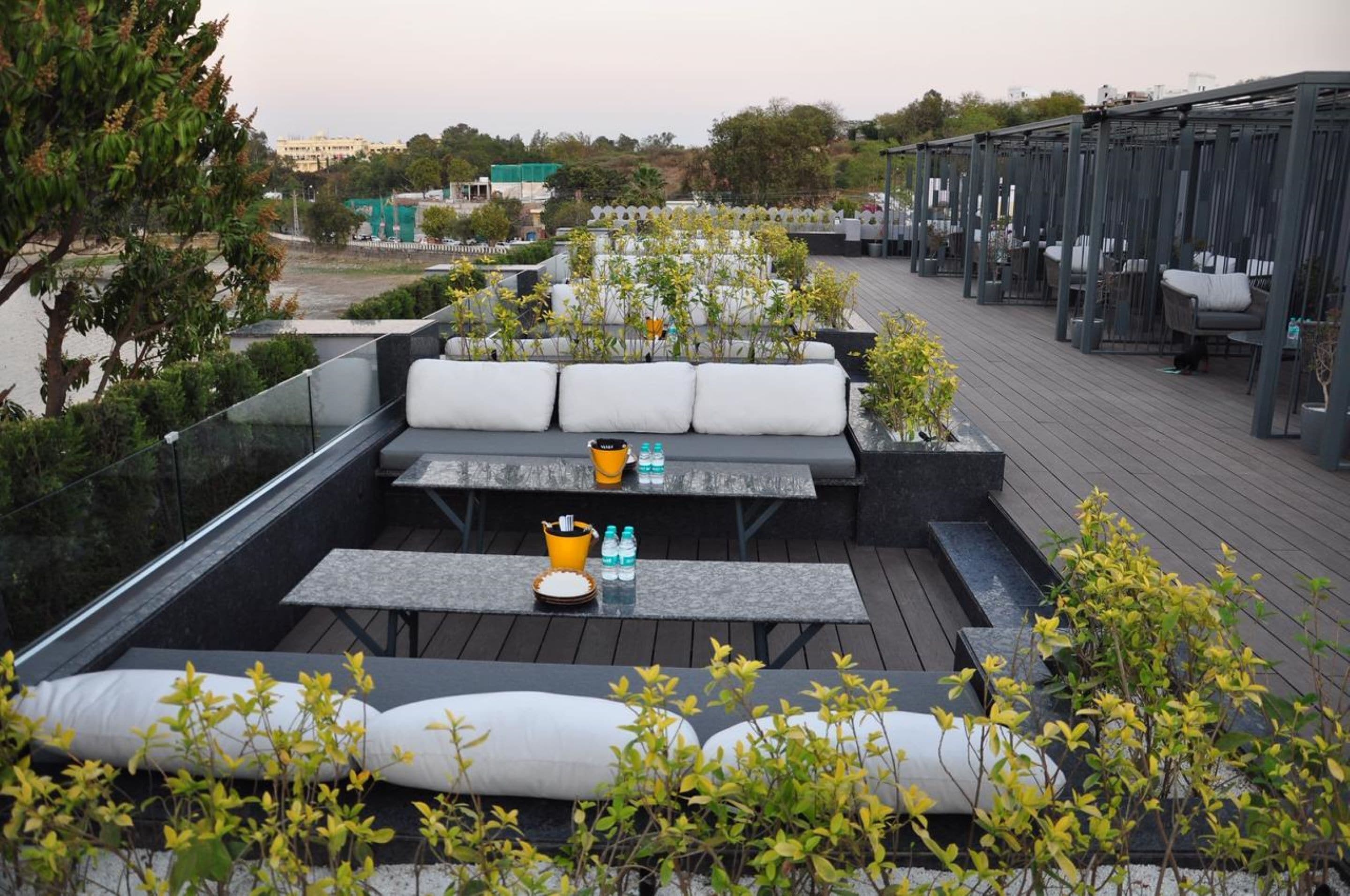 An outdoor seating area with tables and cushioned benches, surrounded by greenery and overlooking a cityscape.