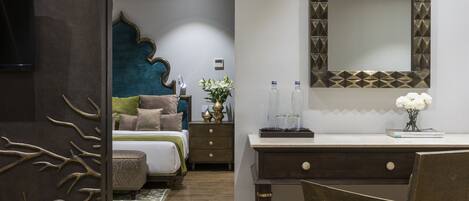A modern bedroom with a wooden floor, a desk with a chair, a bed with a blue headboard, and a mirror with a geometric frame.
