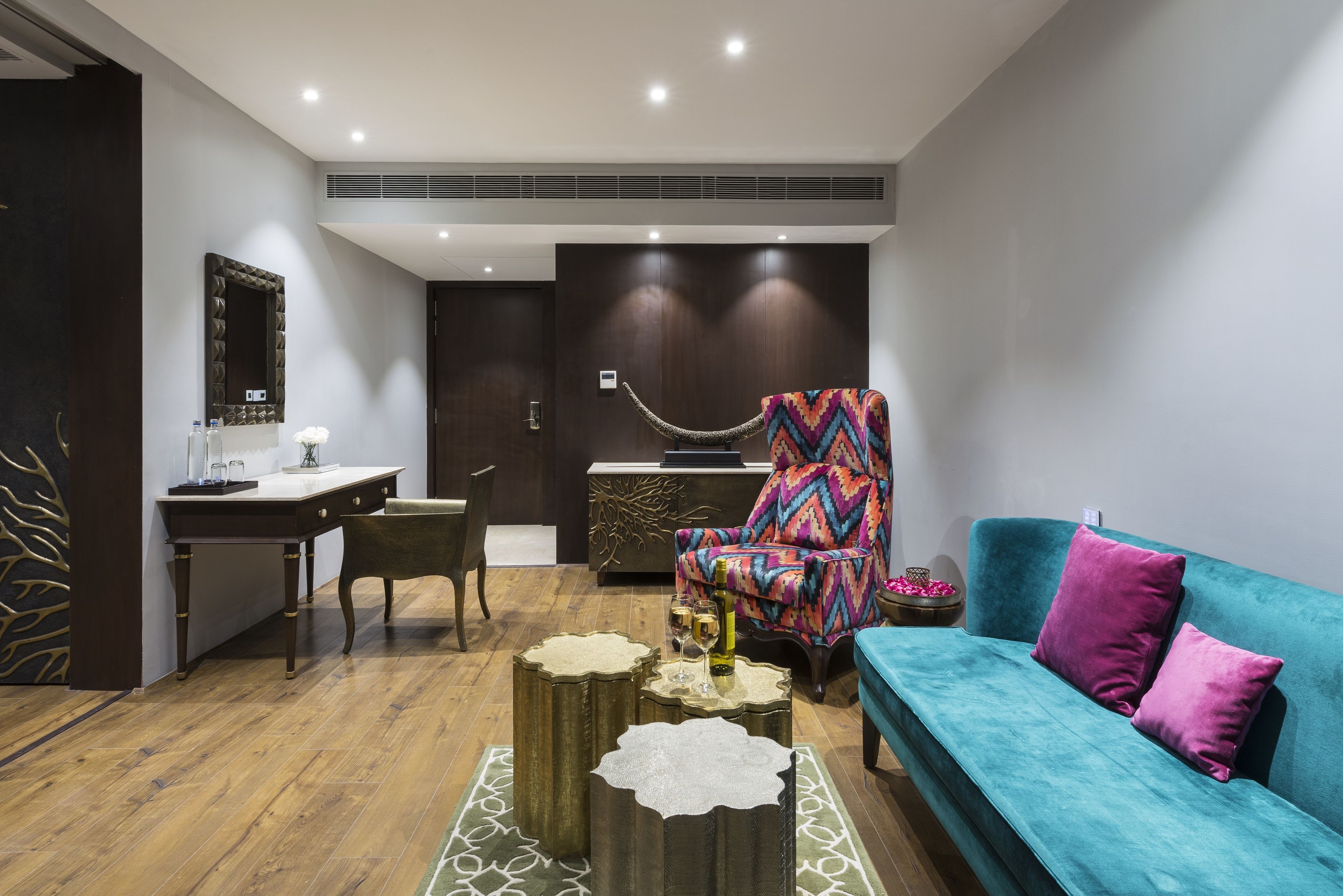 Een moderne woonkamer met een houten vloer, een blauwgroene bank, een kleurrijke fauteuil, een bureau met stoel en een decoratieve spiegel.