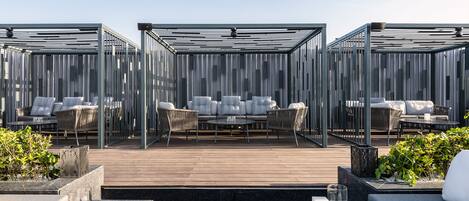 Terraza en la azotea con cubierta de madera, zona de estar exterior con sofás y sillones, y estructura de cerramiento de vidrio.