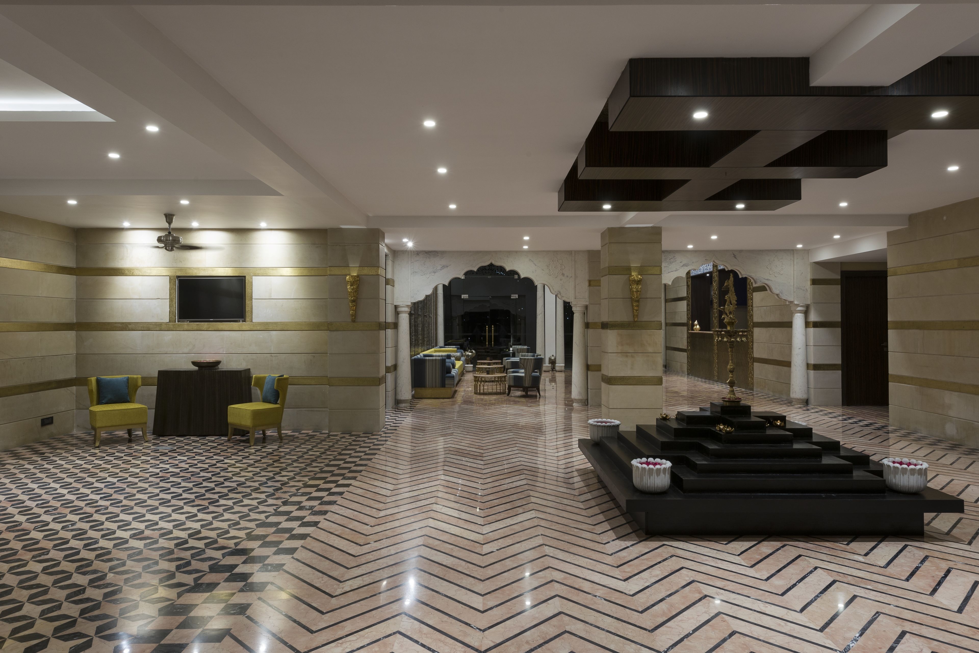 Een ruime lobby met een vloer met geometrisch patroon, een zithoek met gele stoelen, en een centrale decoratieve constructie.