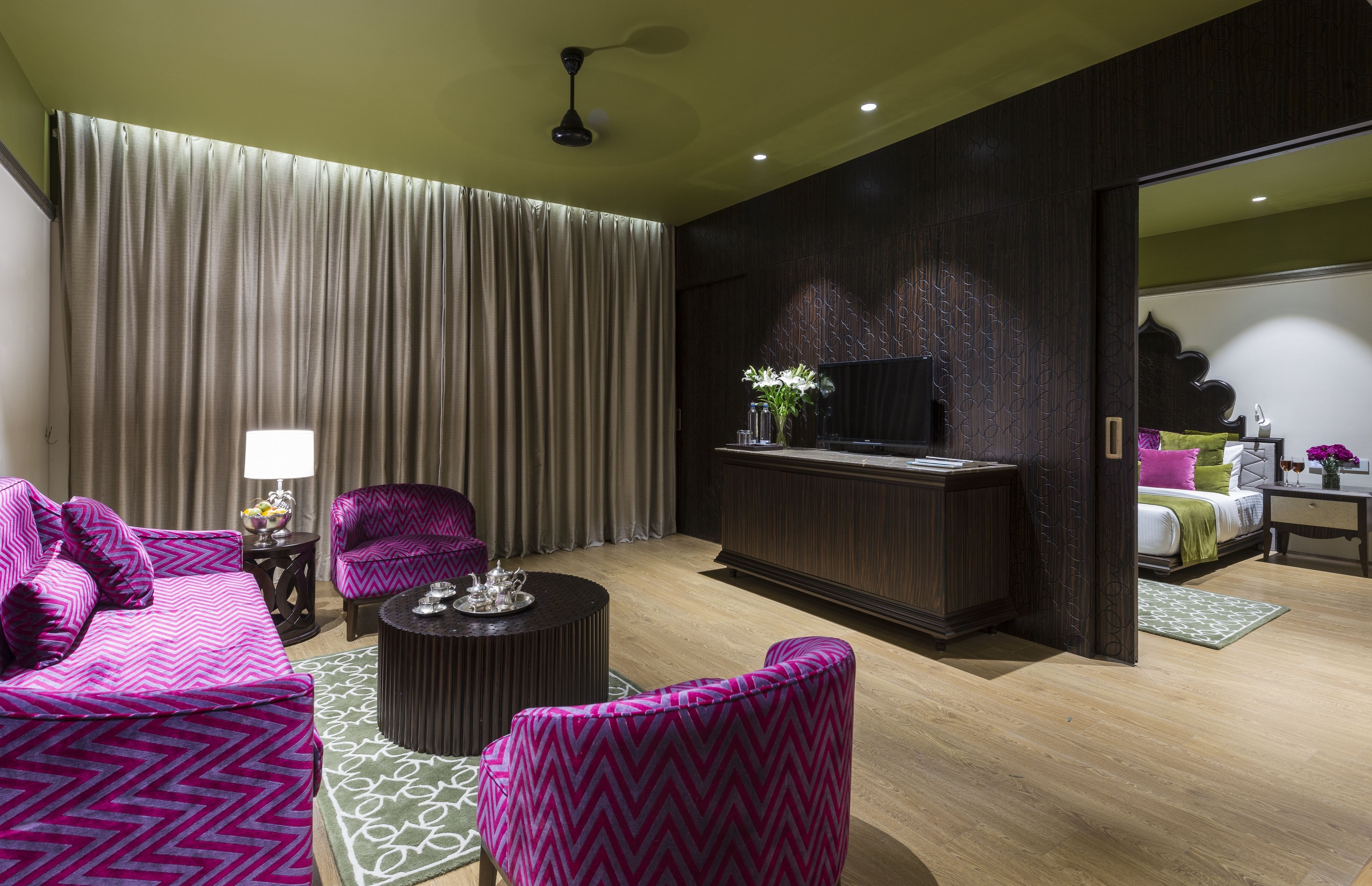 Una habitación de hotel moderna con una cama grande, un área de descanso con sofá y sillones, una mesita redonda pequeña, televisión y un panel de pared de madera oscura.