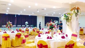 A banquet hall with round tables set for a formal event, decorated with floral arrangements and yellow chairs with red sashes. - Travellers Hotel Phinisi (Makassar)