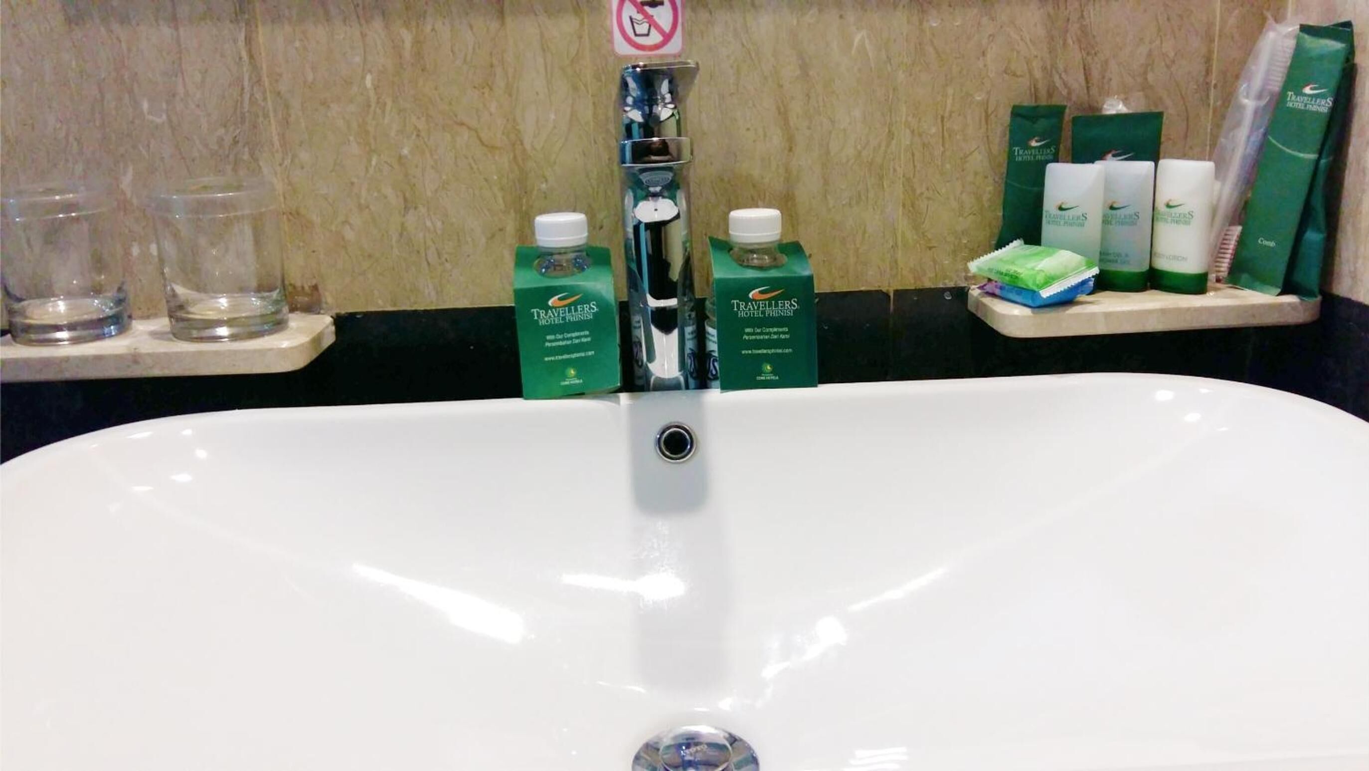 Un lavamanos blanco con grifo cromado, dos dispensadores de artículos de tocador verdes y un cartel de prohibido fumar sobre el lavamanos.