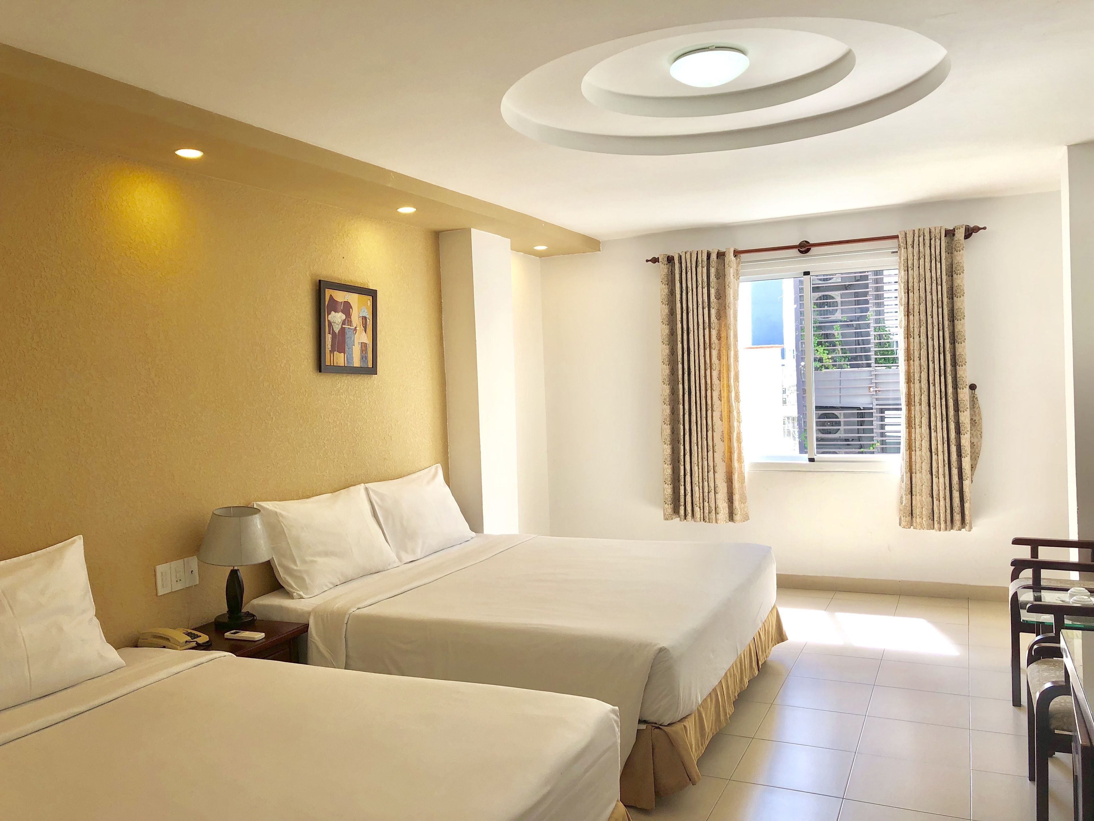 A hotel room with two beds, a desk, a chair, a window with curtains, a ceiling light, and a wall-mounted picture.