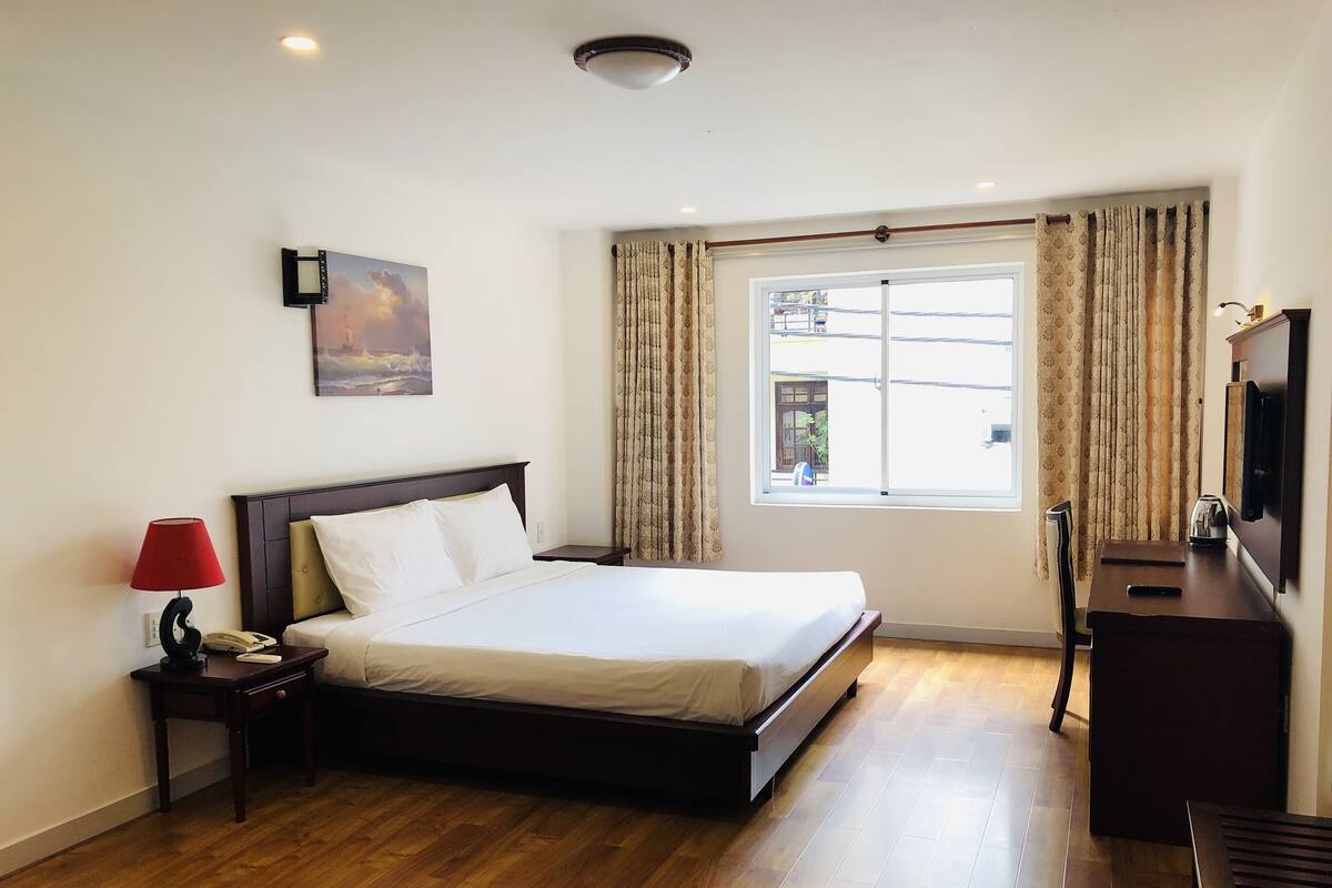 A hotel room with a large bed, a bedside table with a lamp, a desk with a chair, a TV, and a window with curtains.