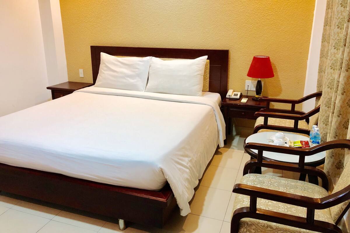 A hotel room with a large bed, a bedside table with a lamp, a chair, and a small table with a teapot and cups.