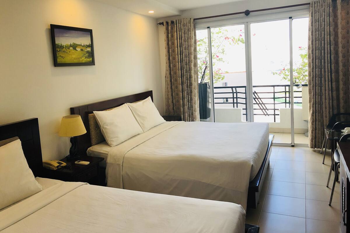 A hotel room with two beds, a nightstand with a lamp, a painting on the wall, and a balcony with a view of the outside.