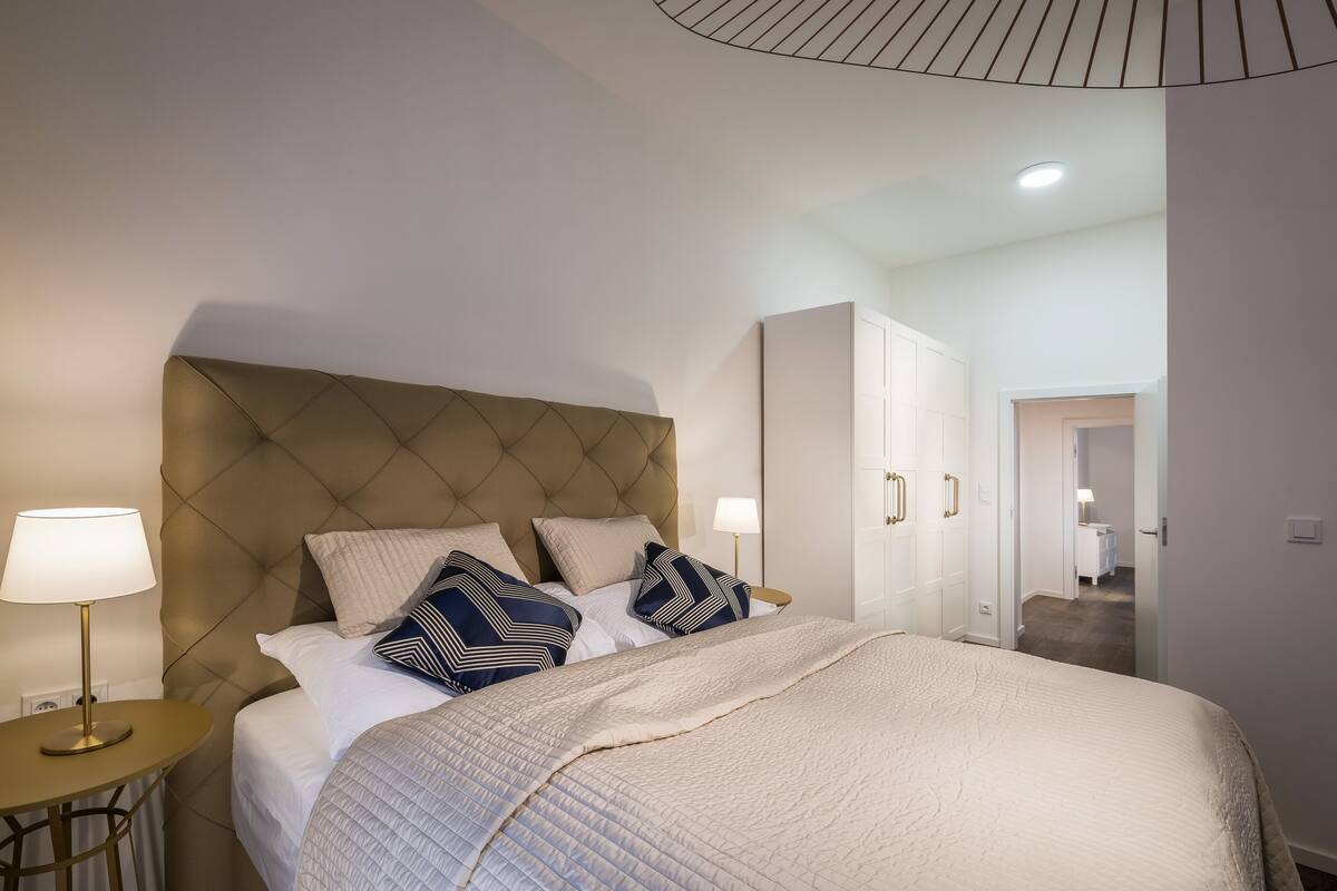 Une chambre à coucher avec une tête de lit capitonnée, un lit avec des oreillers blancs et à motifs, une table de chevet avec une lampe et une armoire.