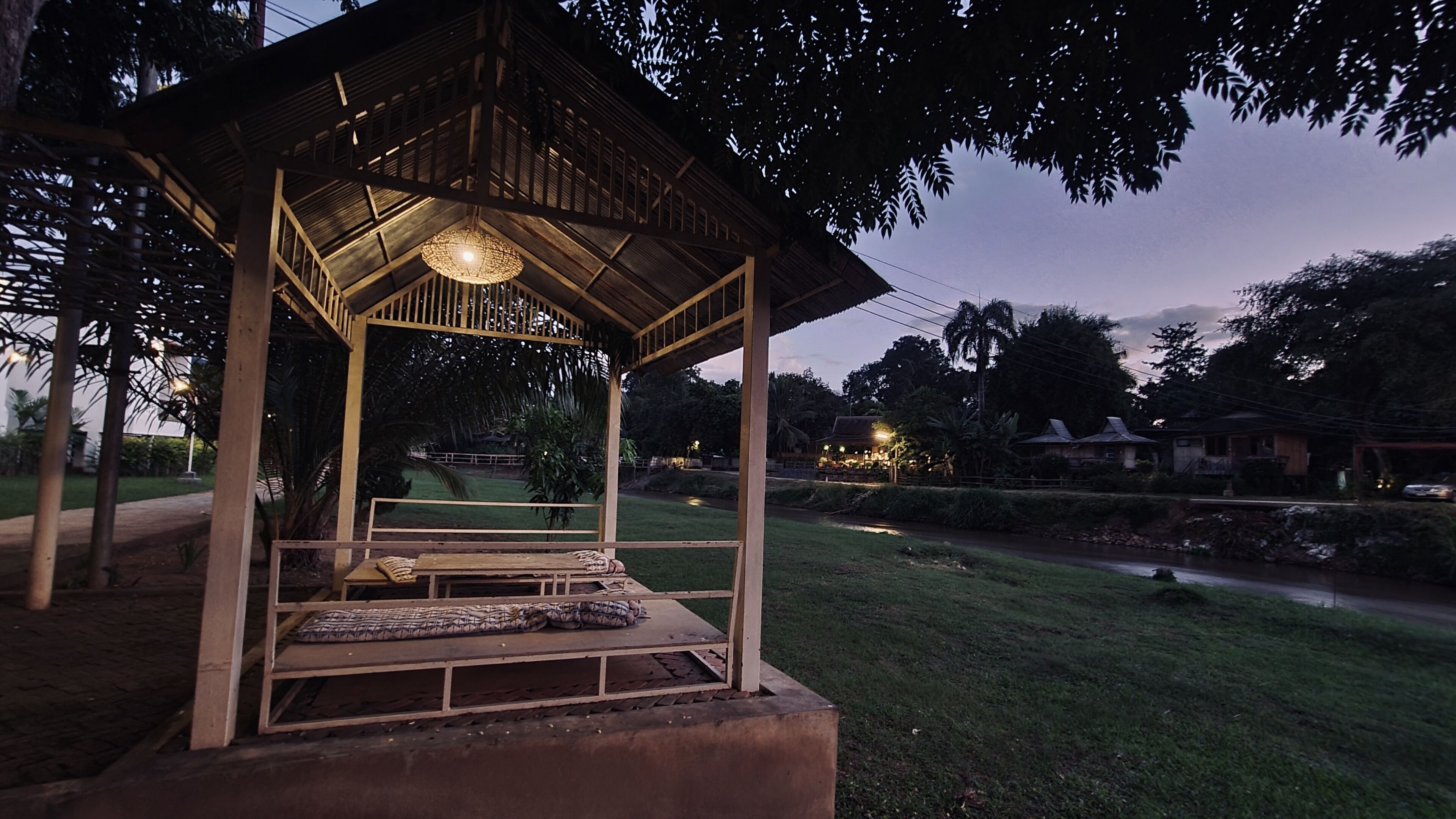 A wooden gazebo with a bench and a hanging light fixture, surrounded by greenery and a pathway.