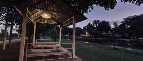 A wooden gazebo with a bench and a hanging light fixture, surrounded by greenery and a pathway.