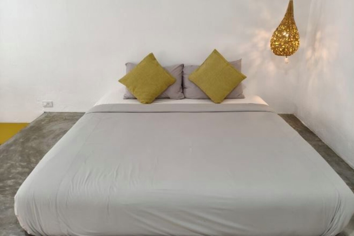 Una cama con ropa de cama blanca y dos almohadas mostaza, una lámpara de pared con textura y una alfombra gris.