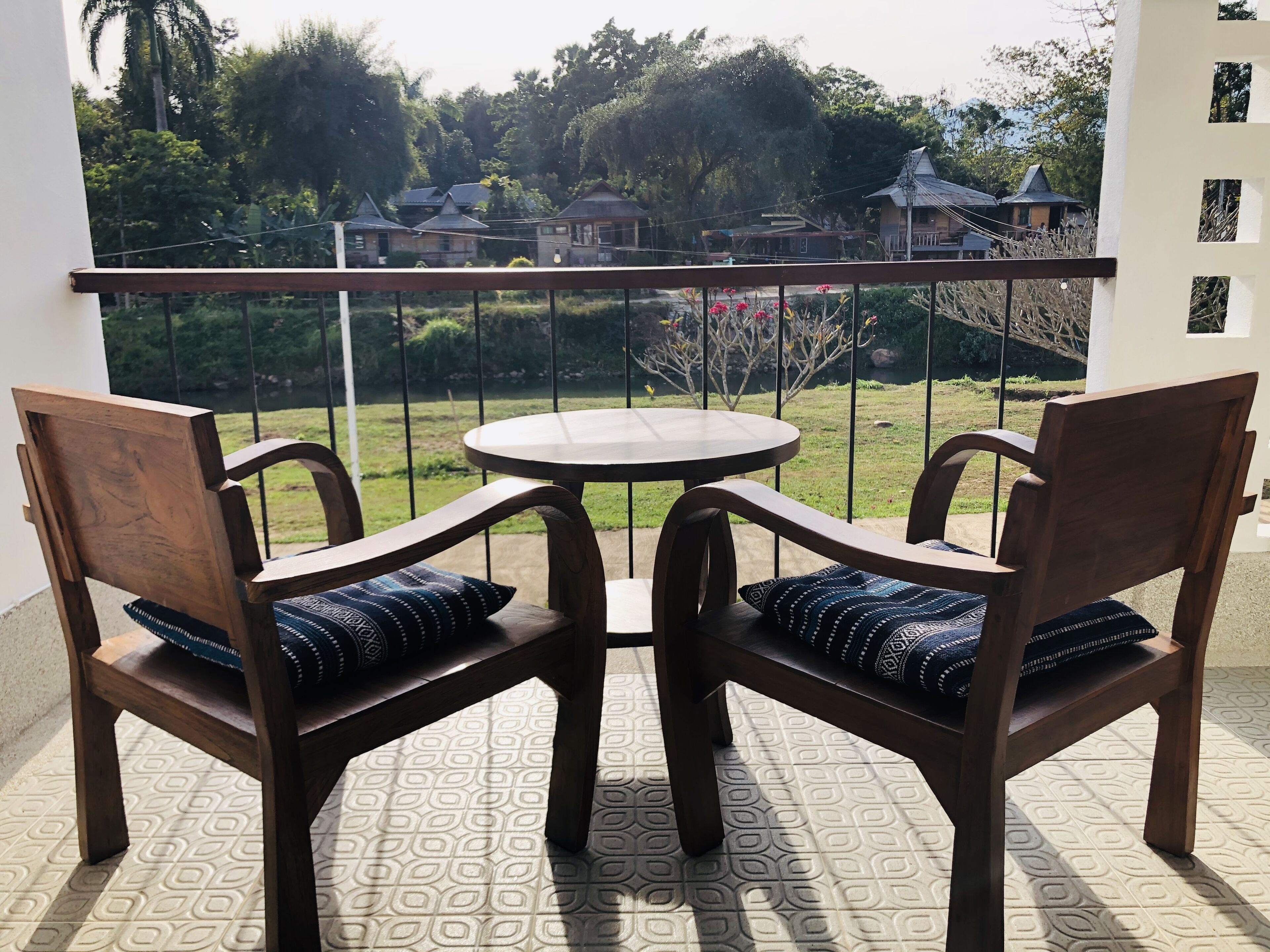 A balcony with two wooden chairs and a small round table, overlooking a garden and distant buildings.