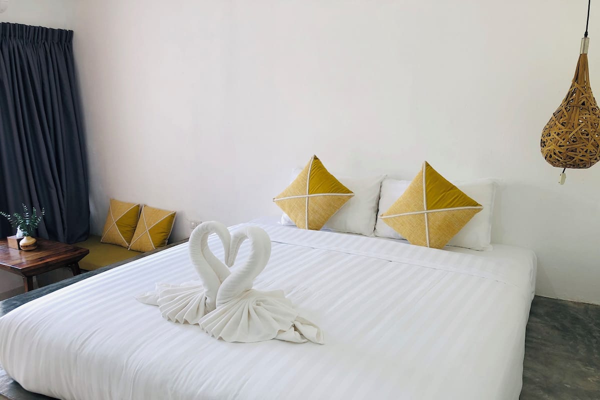 Una cama bien hecha con un arreglo de toallas con forma de cisne, una mesita de noche de madera con una planta y dos almohadas amarillas sobre un colchón blanco.