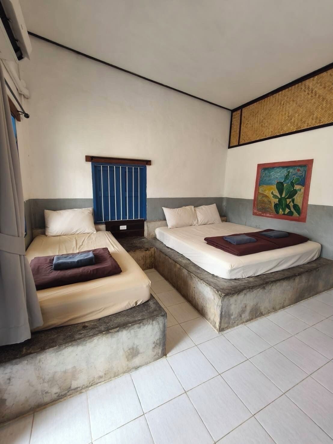 Quarto com duas camas, uma em plataforma baixa e outra elevada. Há uma janela com persianas azuis, um quadro na parede e um chão em azulejos.