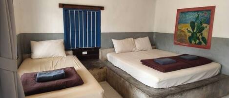 Quarto com duas camas, uma em plataforma baixa e outra elevada. Há uma janela com persianas azuis, um quadro na parede e um chão em azulejos.