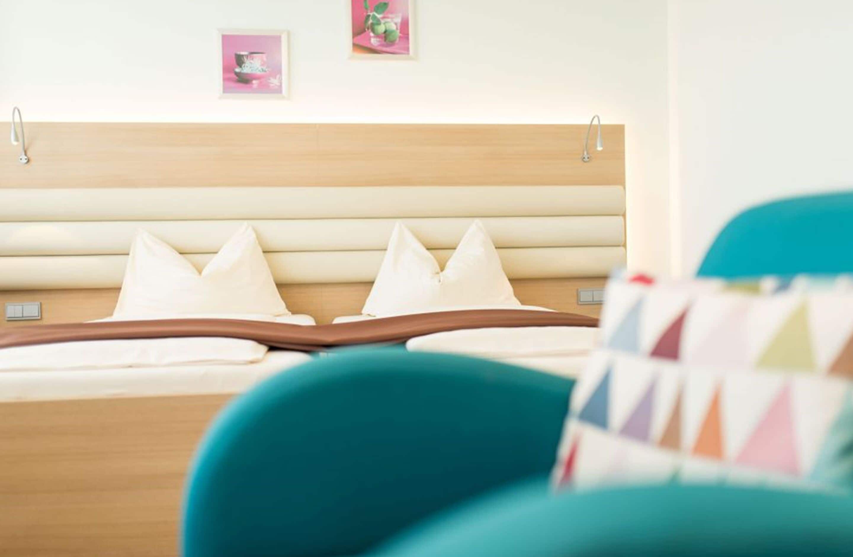 A bed with a wooden headboard, two pillows, and a teal chair in the foreground.