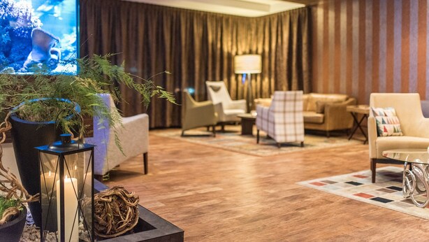 Een moderne hotel lobby met een grote televisie, comfortabele zitplaatsen en een decoratieve plantencompositie.