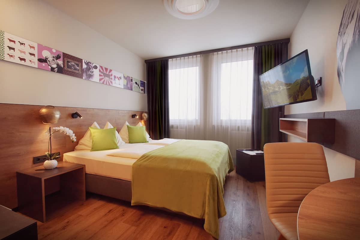 A hotel room with a bed, bedside table with a lamp, a TV mounted on the wall, and a chair.