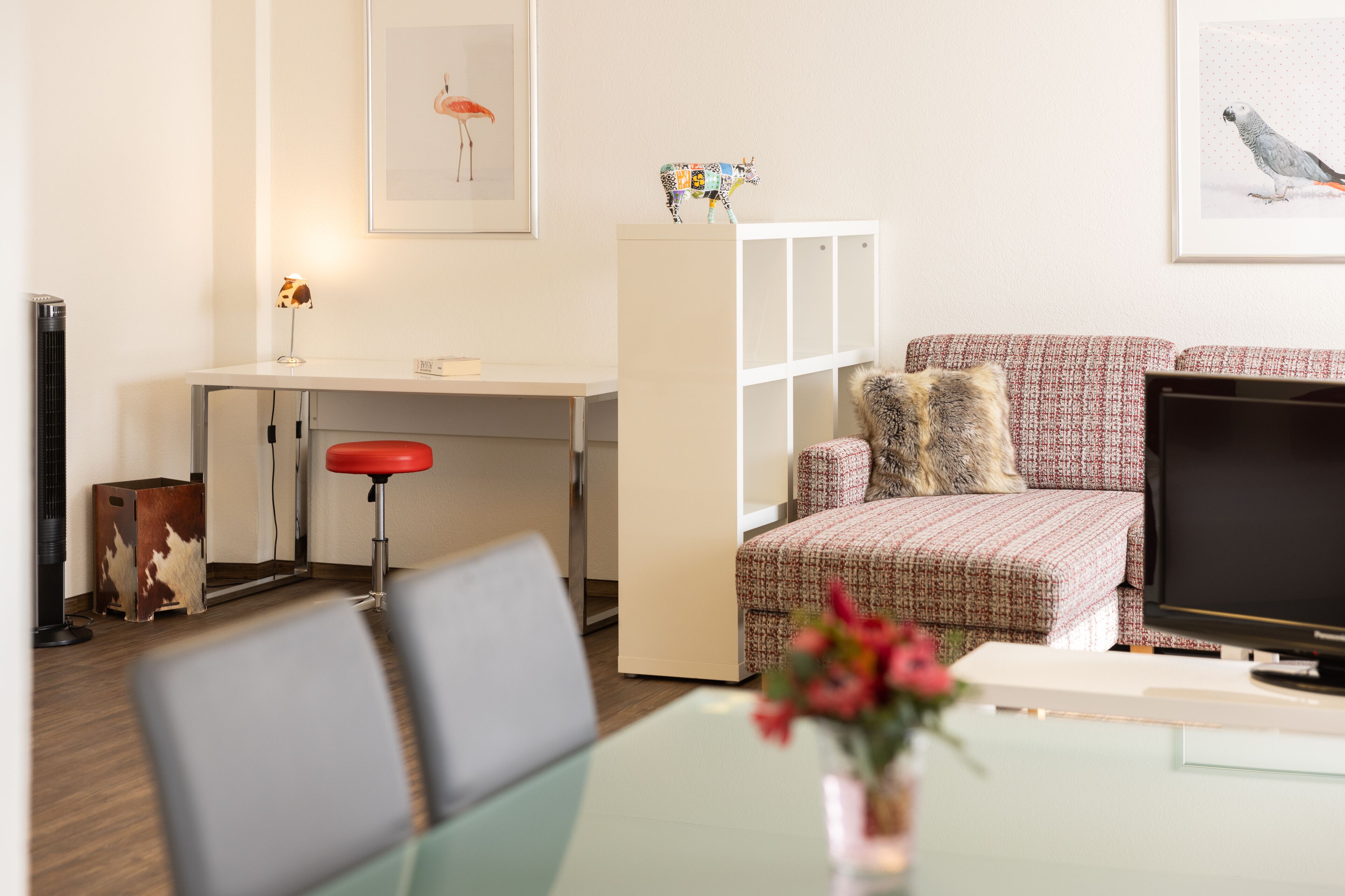 A modern living room with a sofa, a TV, a desk with a chair, and a small table with a vase of flowers.