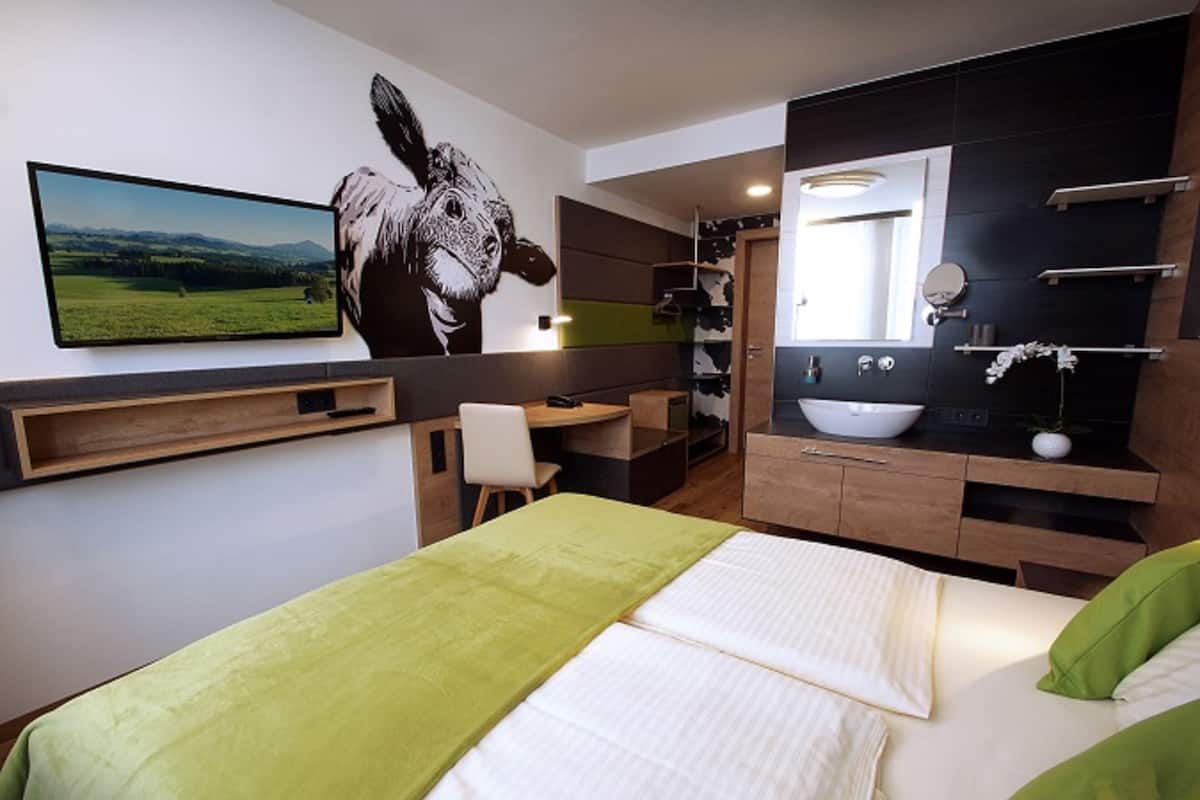 A modern hotel room with a large bed, a desk, a bathroom with a mirror and sink, and a cow mural on the wall.