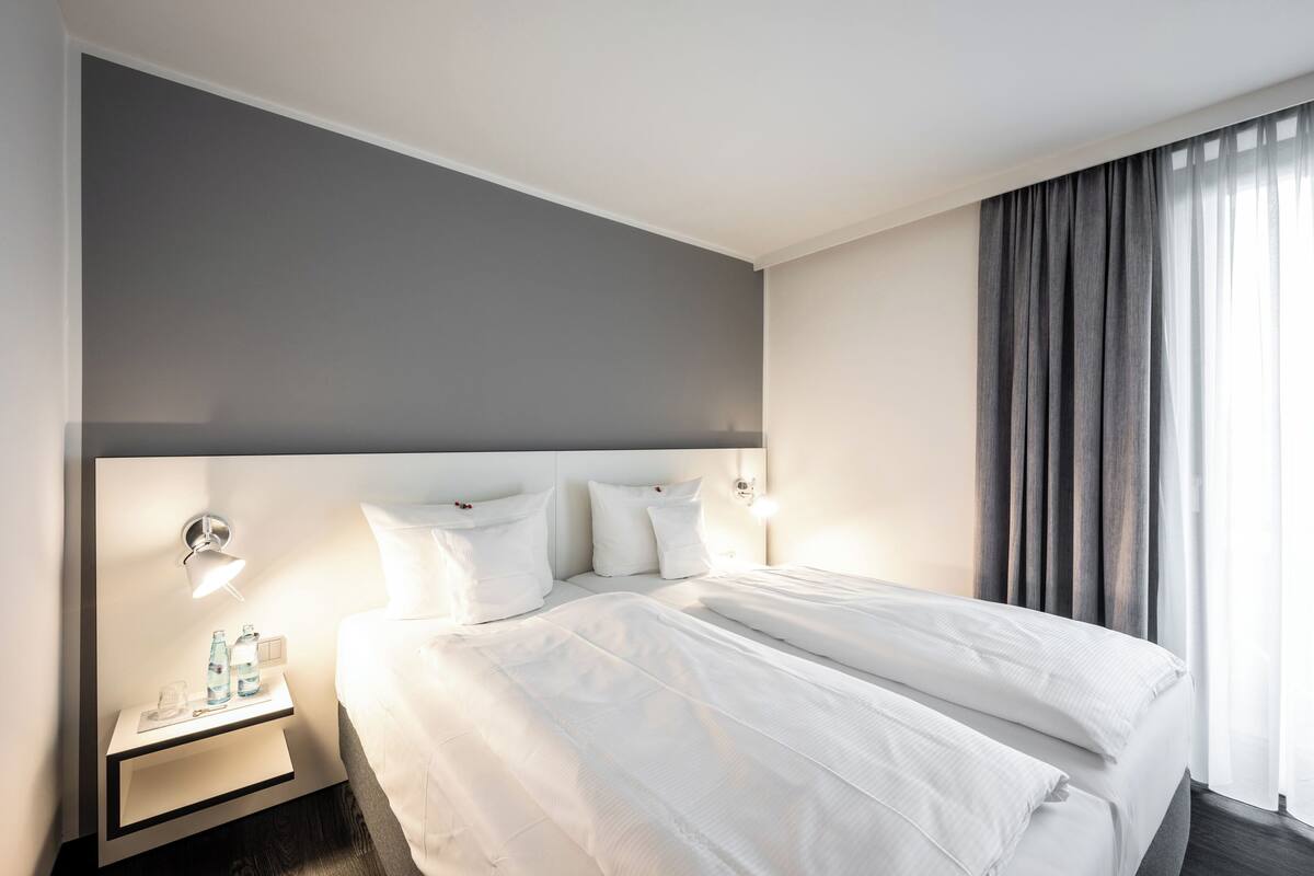 Een hotelkamer met een groot bed, een nachtkastje met een lamp en een raam met gordijnen.