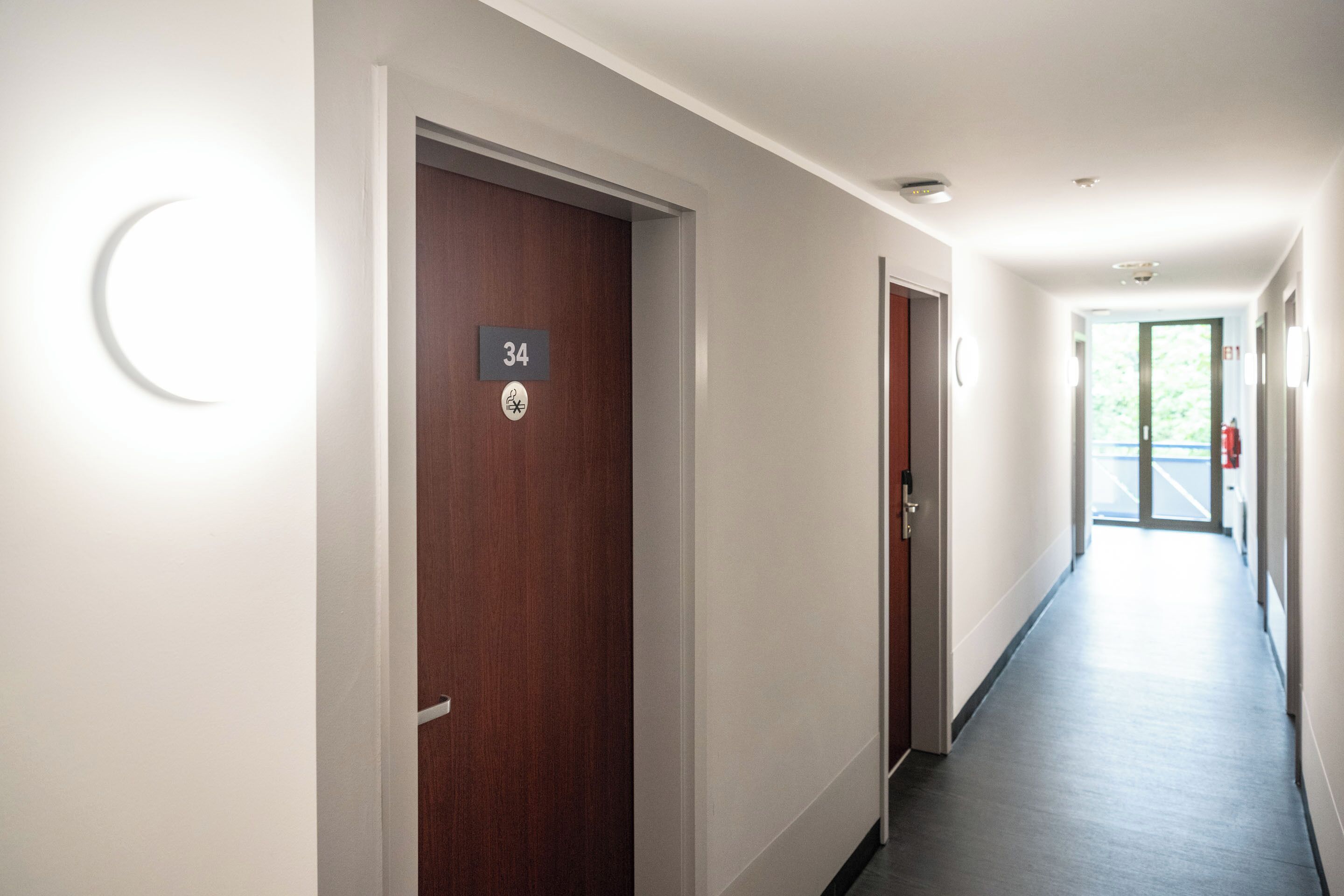 A corridor with a wooden door labeled "34", a white wall with a crescent moon light, and a view of a building outside.