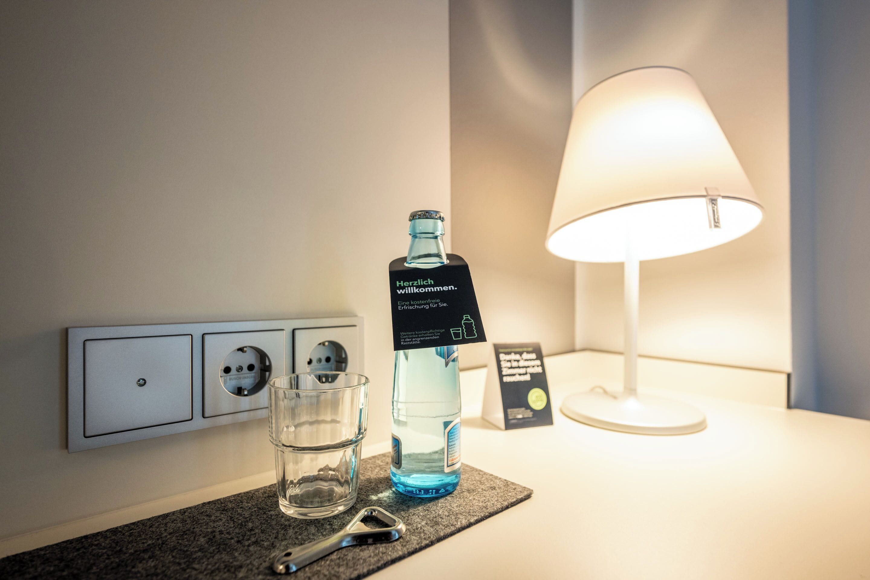A hotel room with a water bottle, glasses, and a lamp on a table.
