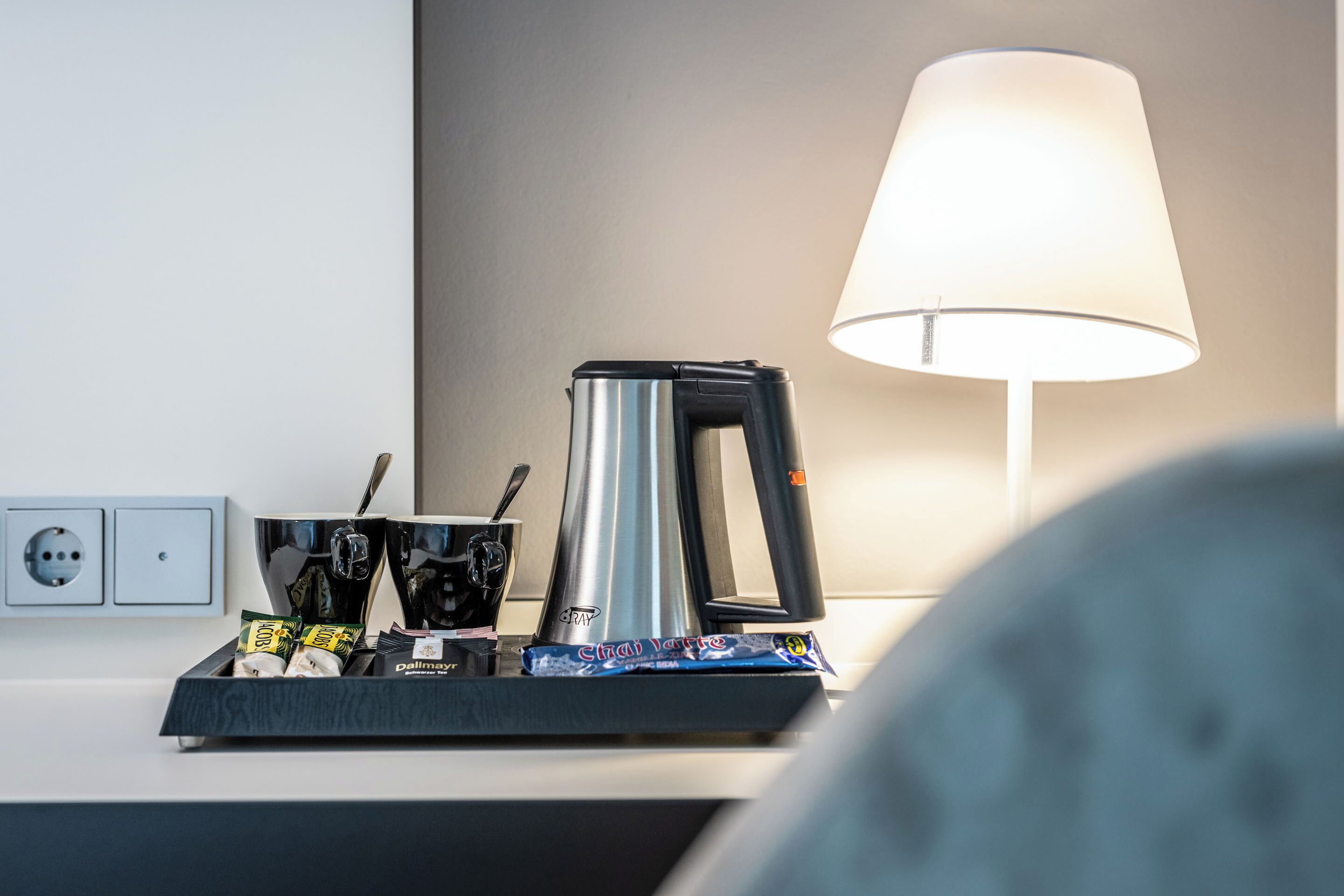 Een koffiezetapparaat, een lamp en een dienblad met koffiekopjes en een chocoladereep op een hotel aanrecht.