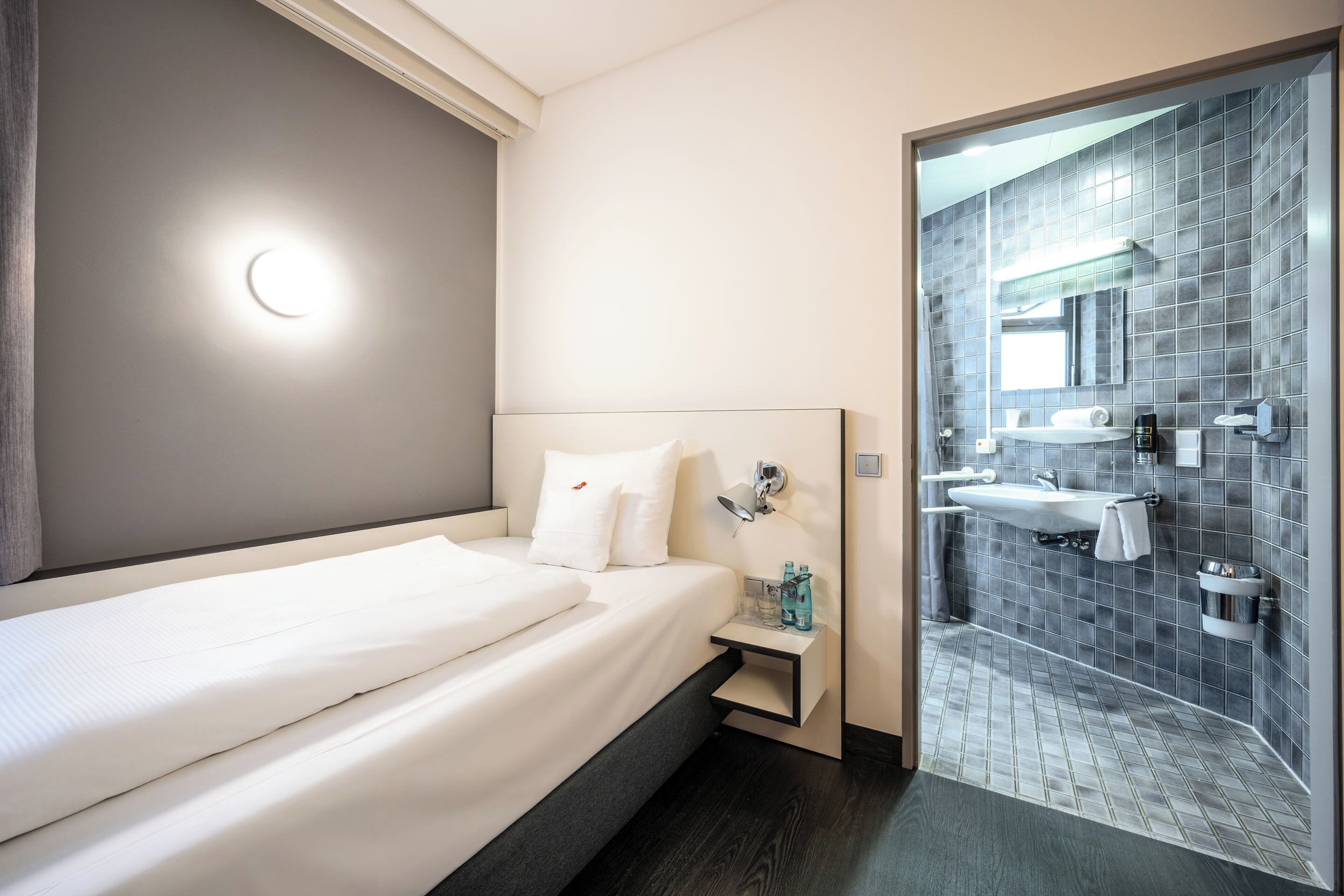 Una habitación de hotel con cama, mesita de noche y baño con lavamanos y espejo.