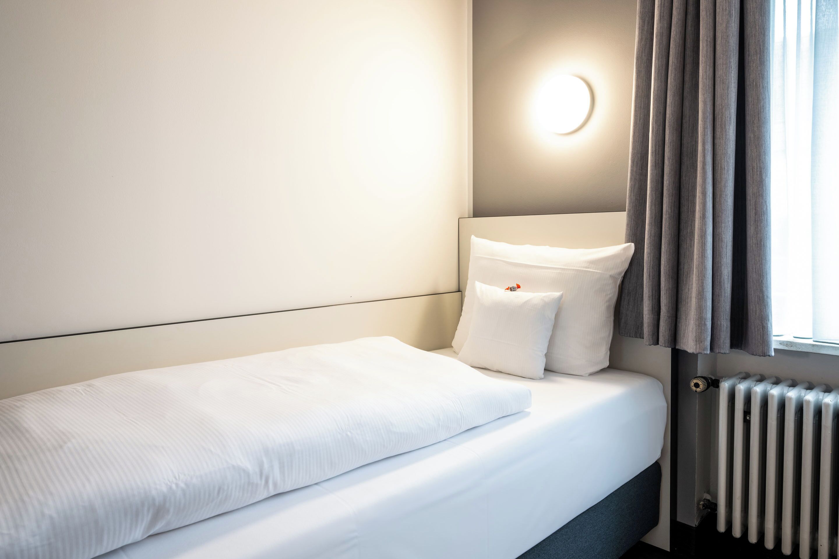 Ein ordentlich bezogenes Bett mit weißen Laken, ein Nachttisch mit Lampe und ein Fenster mit Vorhängen.