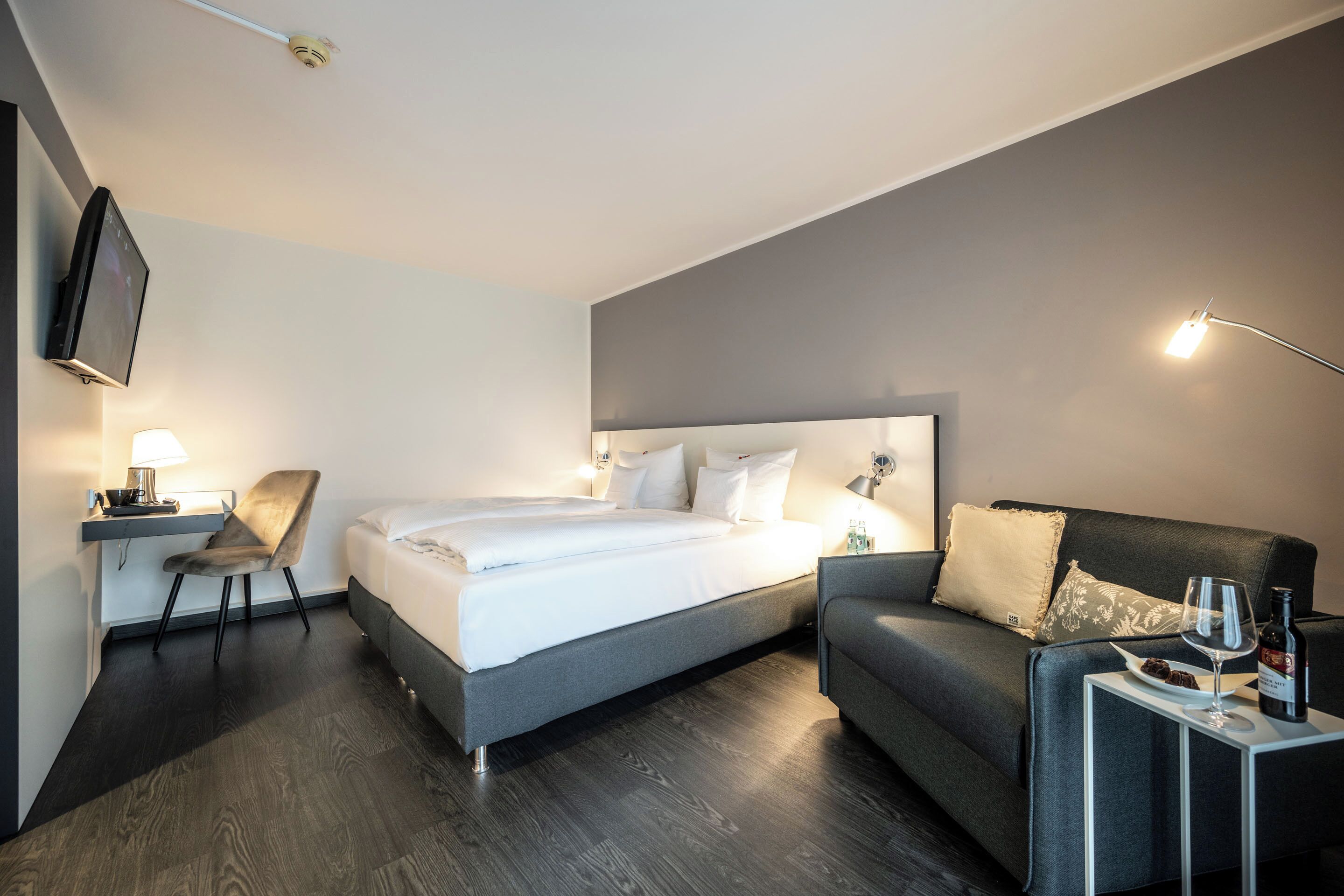 A modern hotel room with a large bed, a sofa, a desk with a chair, a TV, and a small table with a wine glass and bottle.
