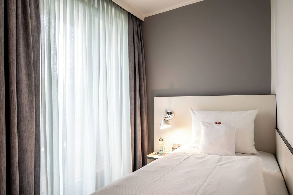 Een hotelkamer met een bed, nachtkastje, lamp en raam met gordijnen.