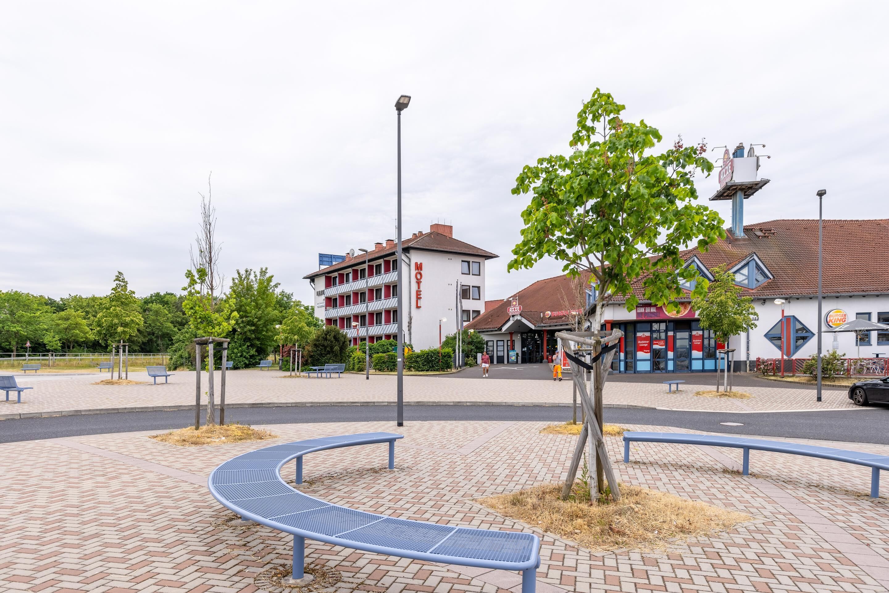 A motel with a red and white facade, a parking area, and a small park with benches and a tree.