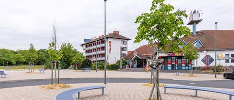Un motel con fachada roja y blanca, una zona de estacionamiento y un pequeño parque con bancos y un árbol.