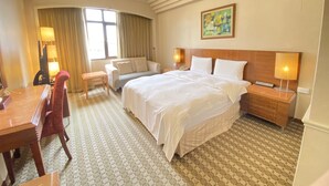 Habitación de hotel con una cama grande, un escritorio, una silla, una mesita, una mesita de noche, una lámpara, un cuadro en la pared y una ventana con cortinas.