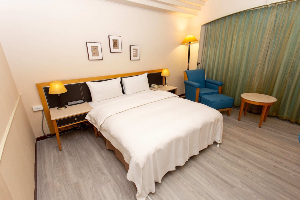 A hotel room with a bed, bedside tables, a desk, a chair, a small round table, and a curtain.