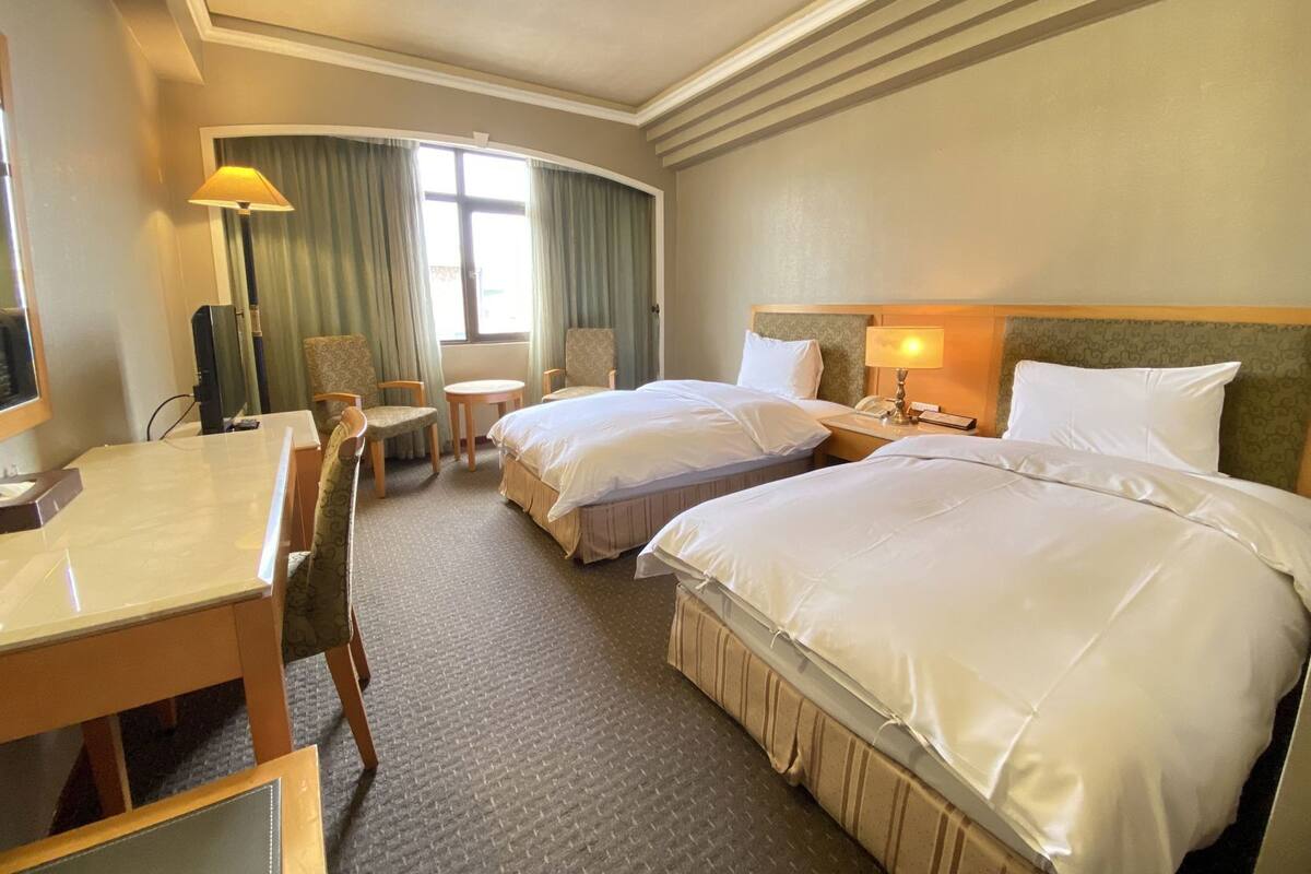 A hotel room with two beds, a desk, a chair, a lamp, and a TV.