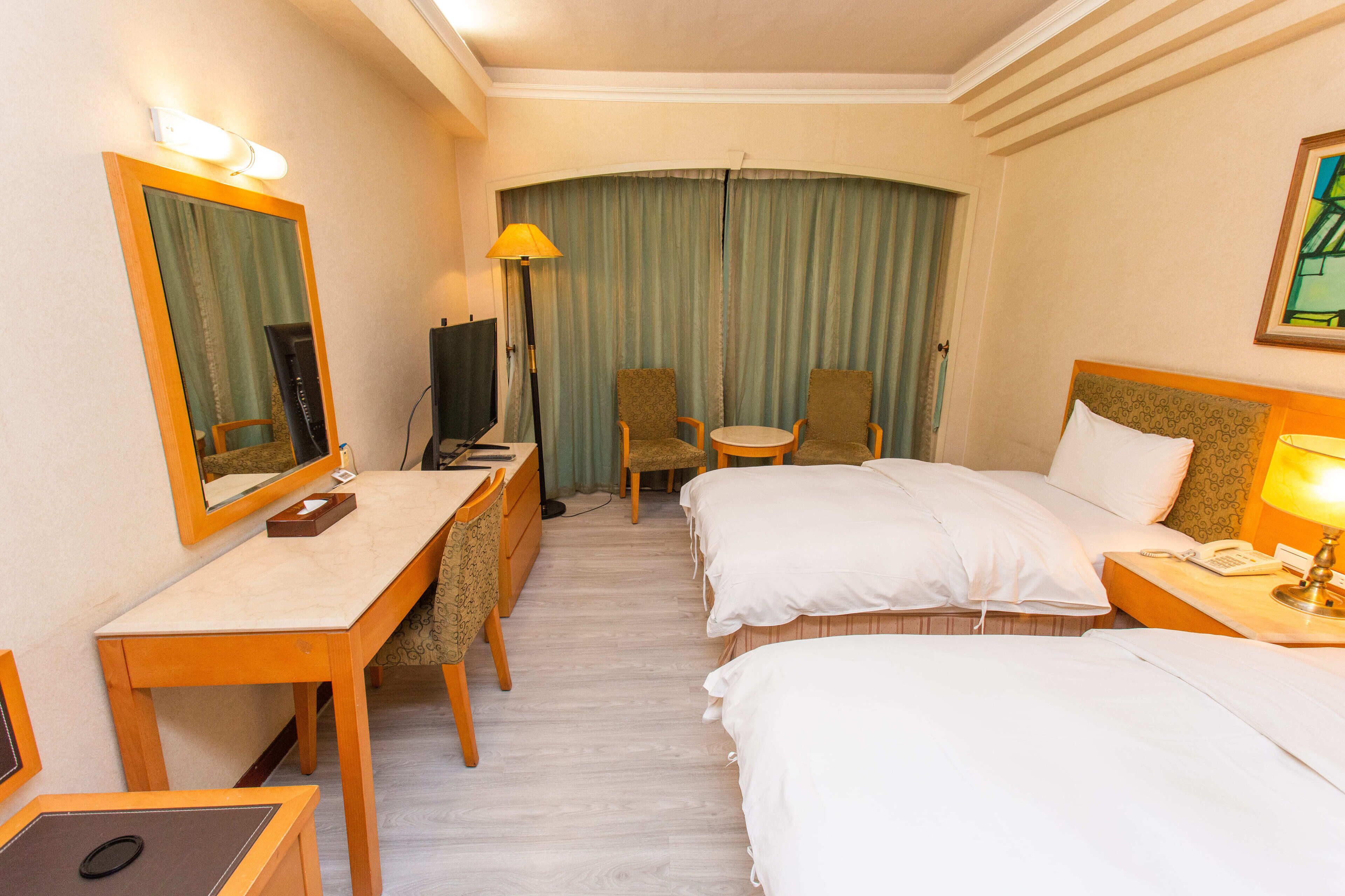 A hotel room with two beds, a desk, a chair, a TV, and a lamp.