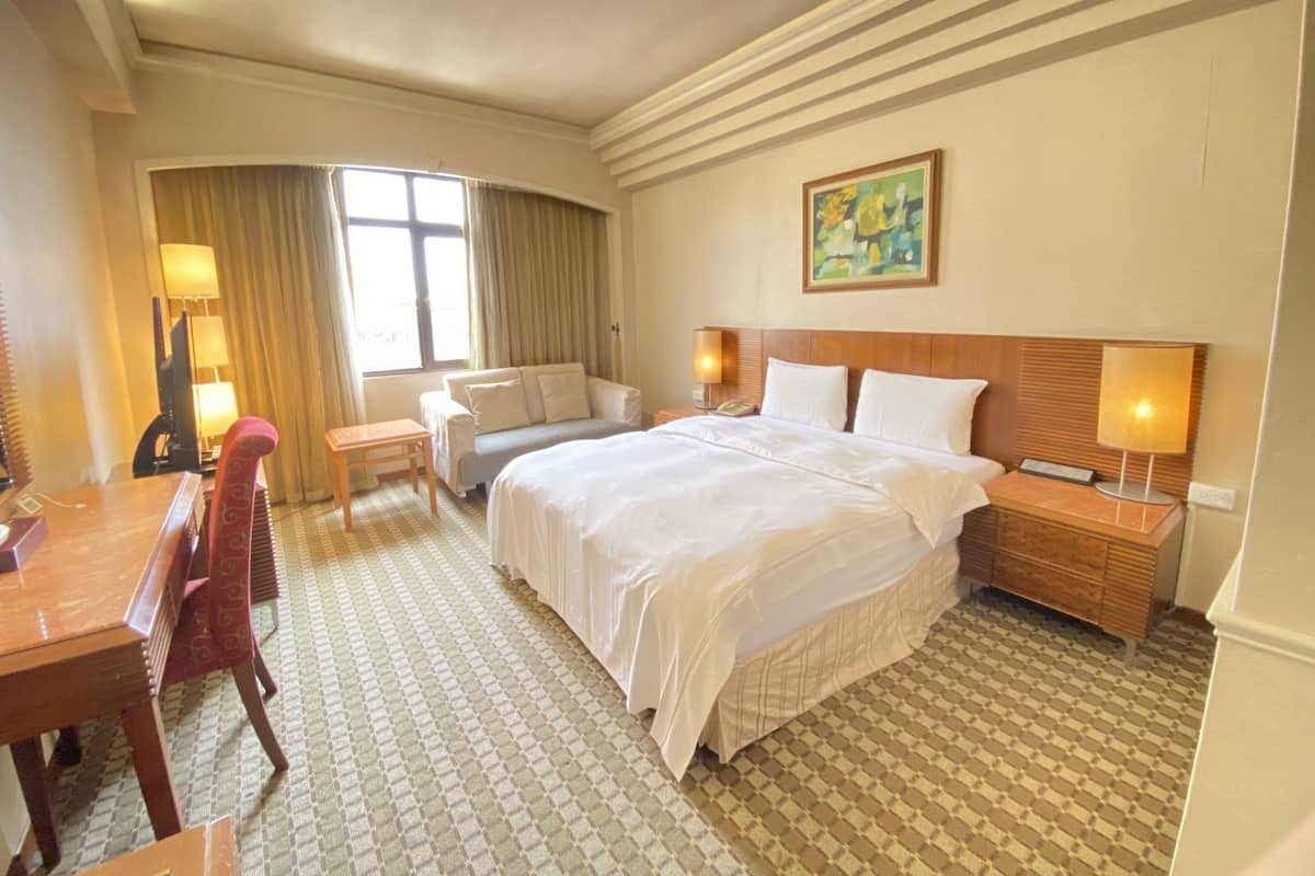 A hotel room with a large bed, a desk, a chair, a sofa, a nightstand, and a painting on the wall.